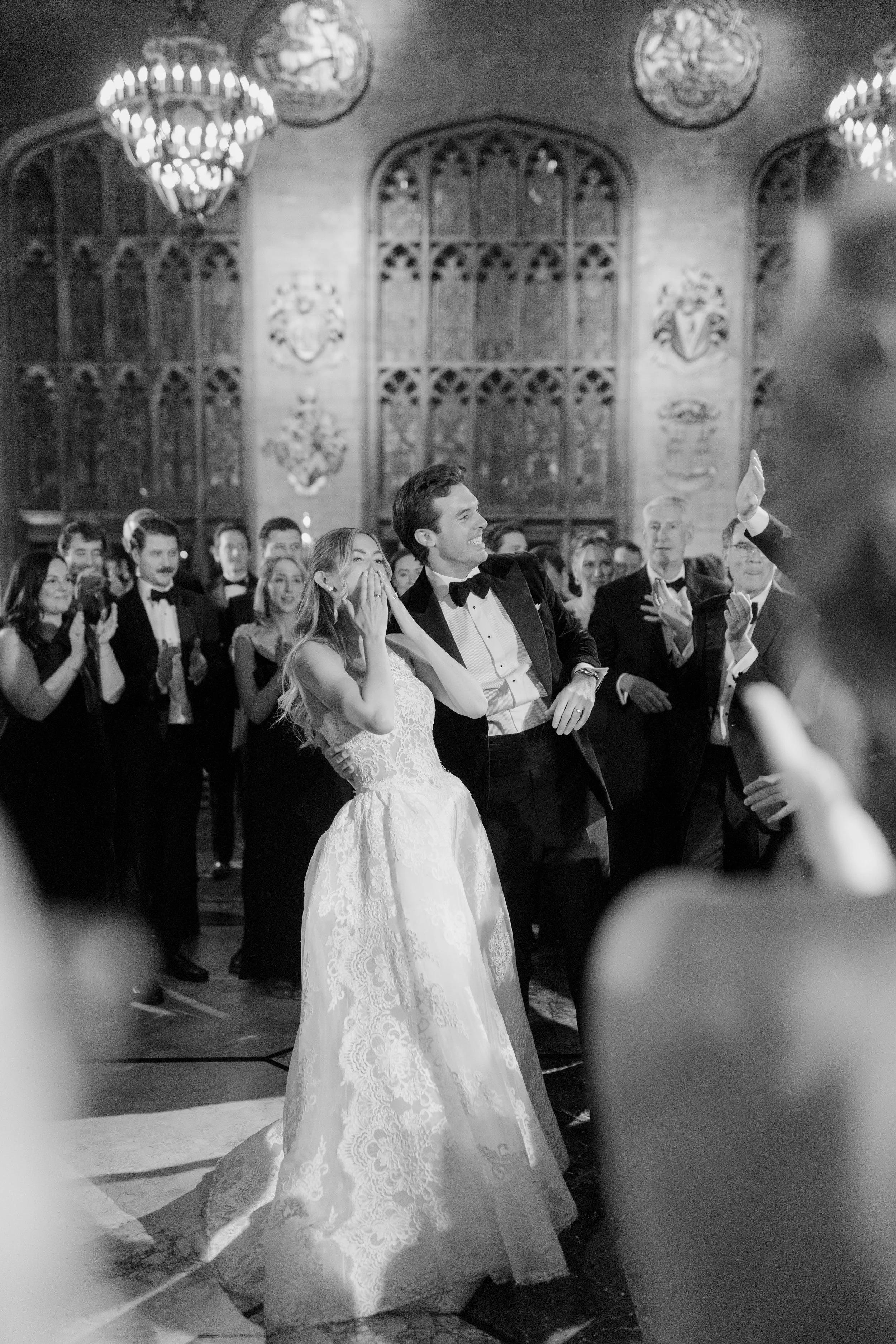 A wedding reception with a bride and groom in the center, surrounded by guests clapping and smiling, in a decorated hall with chandeliers and ornate wall details.