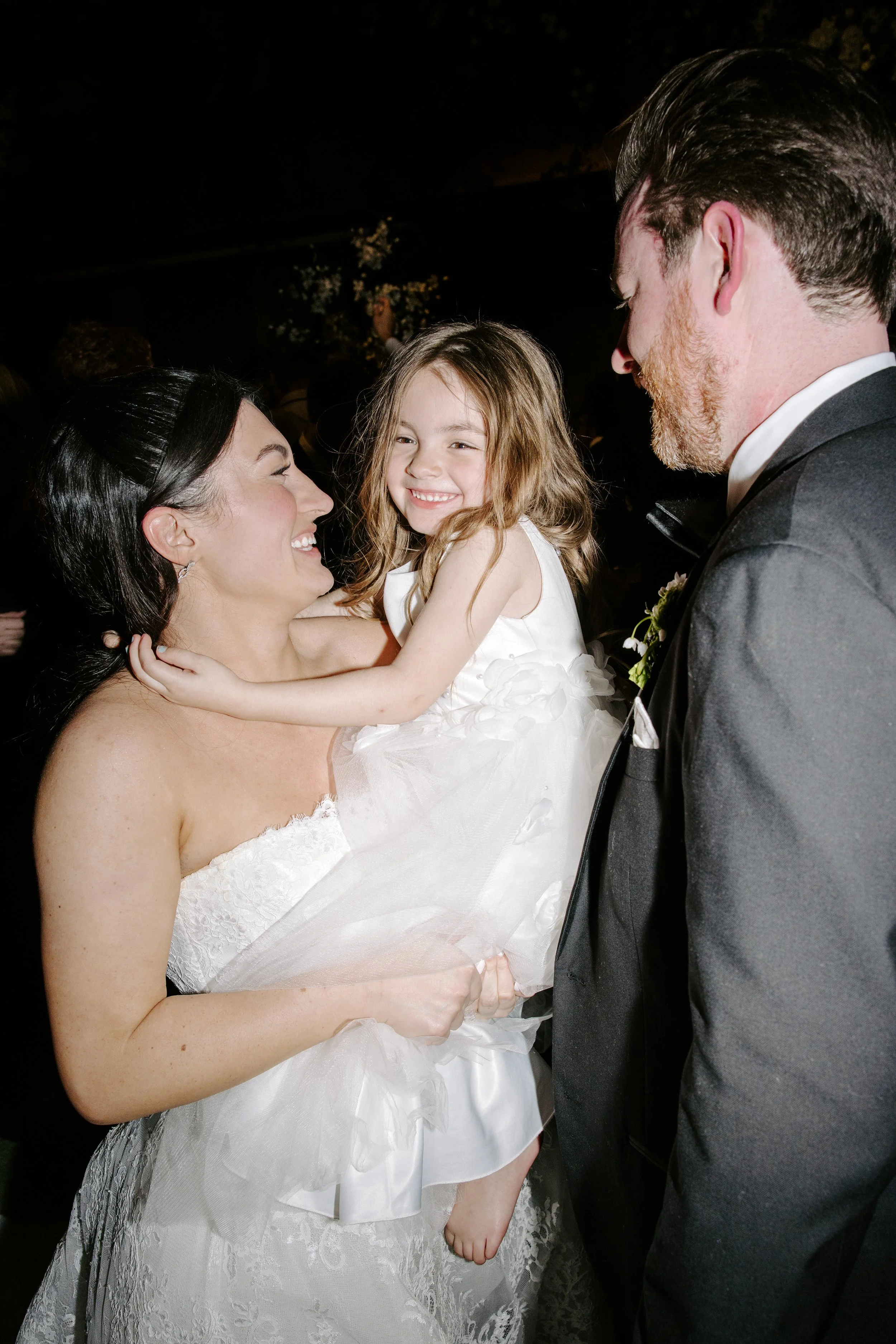 A woman in a wedding dress holding a smiling young girl in a white dress, standing close to a man in a tuxedo at a wedding reception or celebration.