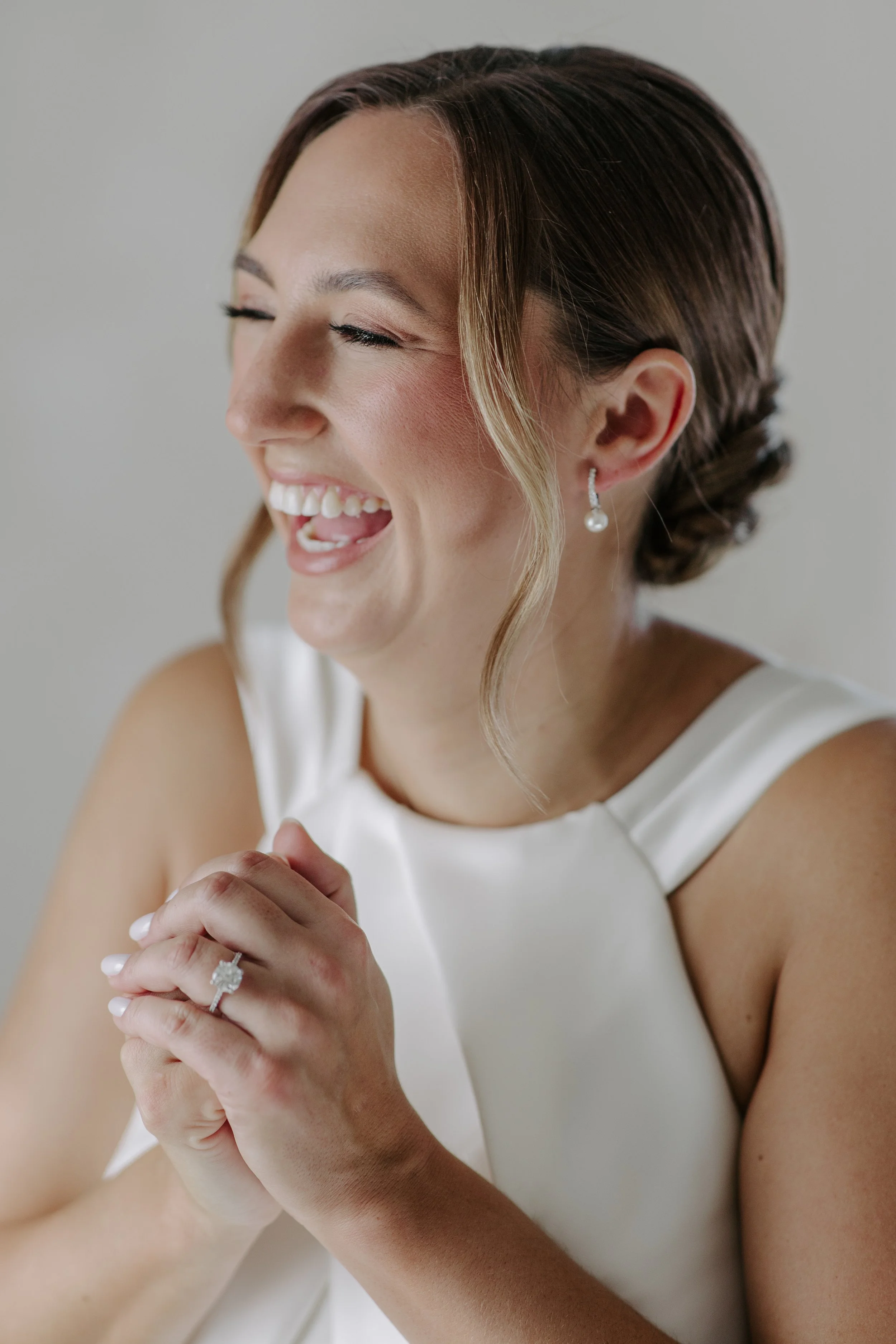 A woman with fair skin, brown hair styled in an elegant updo, is smiling and laughing, with her eyes closed. She is wearing a white dress, pearl earrings, and a diamond engagement ring, and has her hands clasped together.