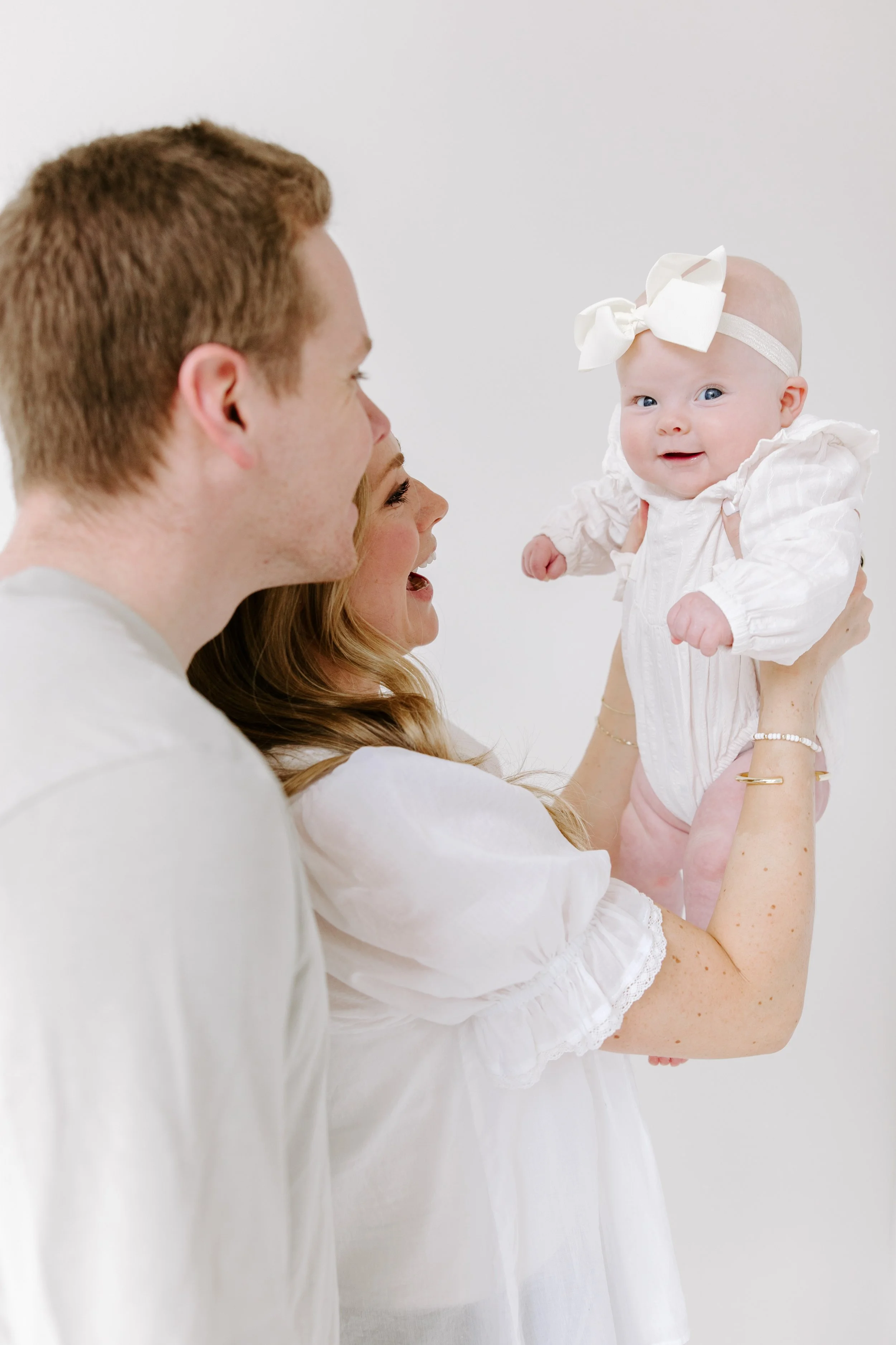A woman holding a smiling baby girl with a large white bow headband against a plain white background.