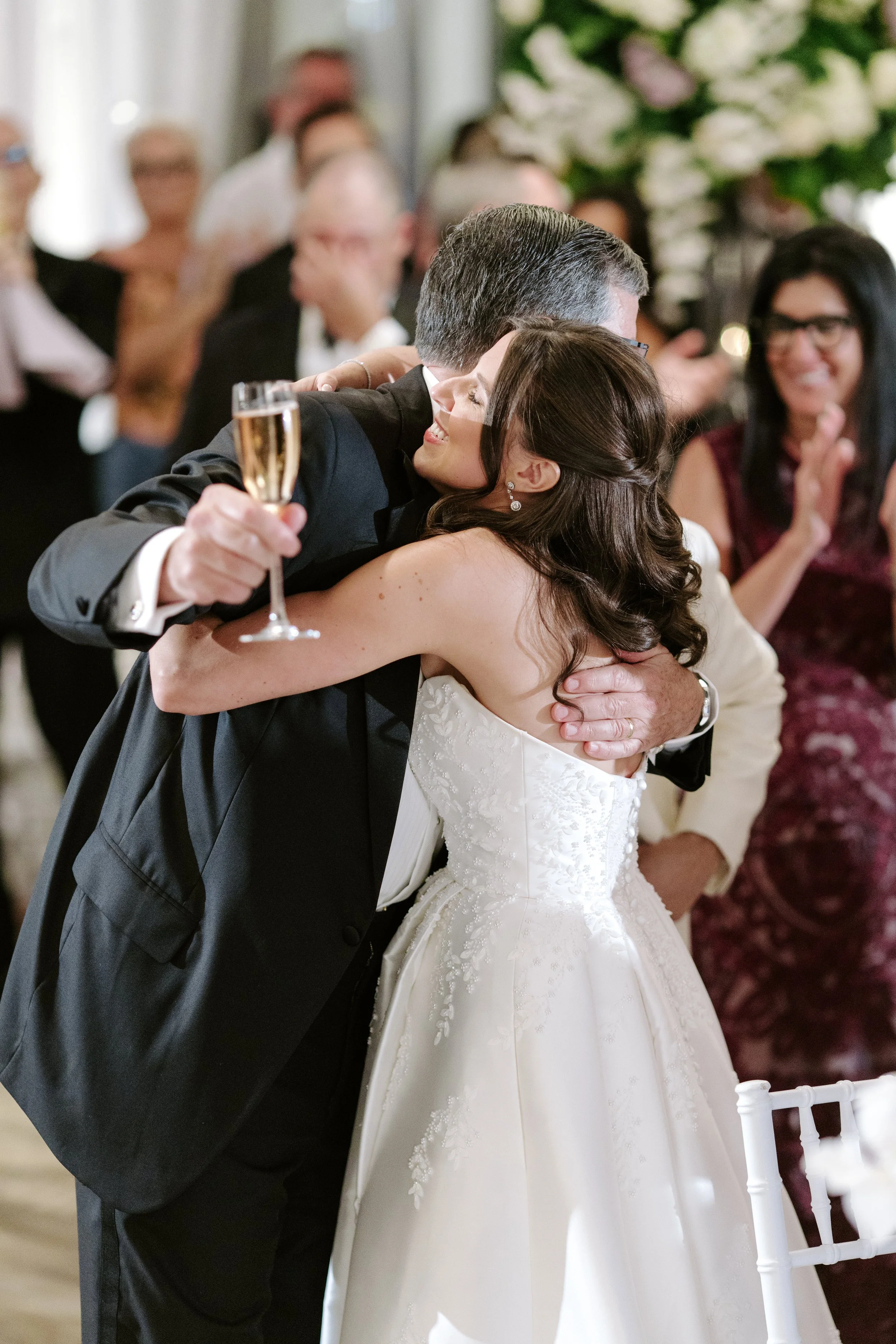 A bride and groom embrace at their wedding reception, the groom holding a glass of champagne, with guests smiling and clapping in the background.