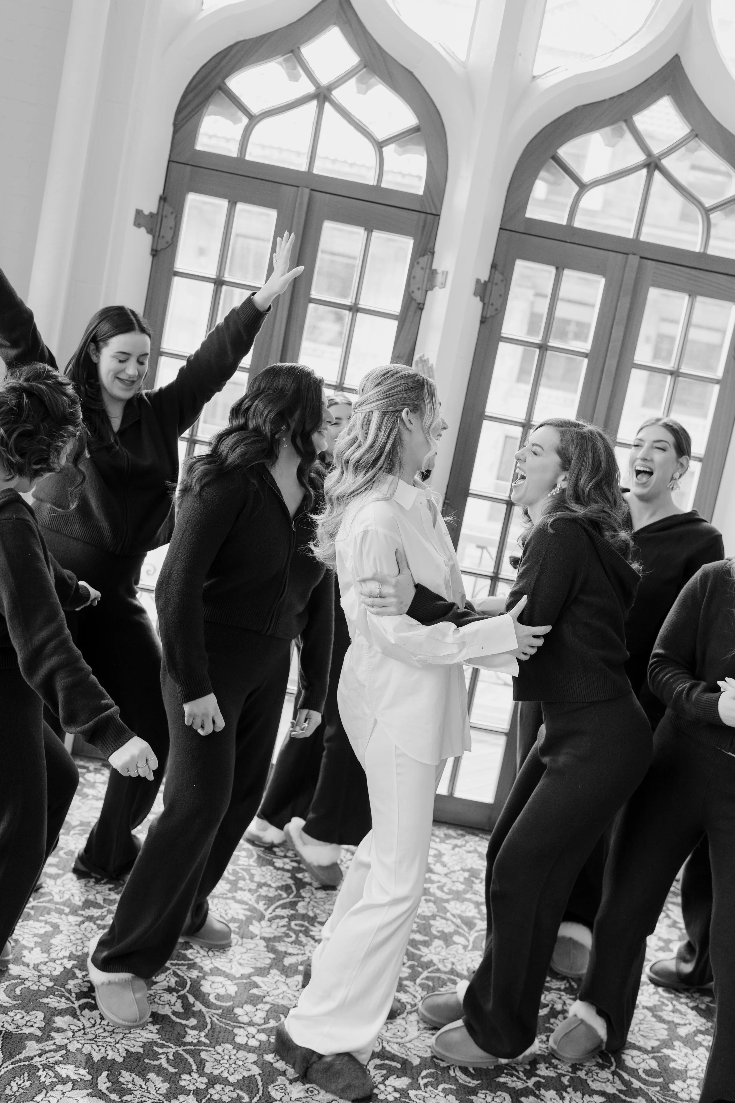 Group of women dancing and laughing indoors near large windows with decorative wood frames.