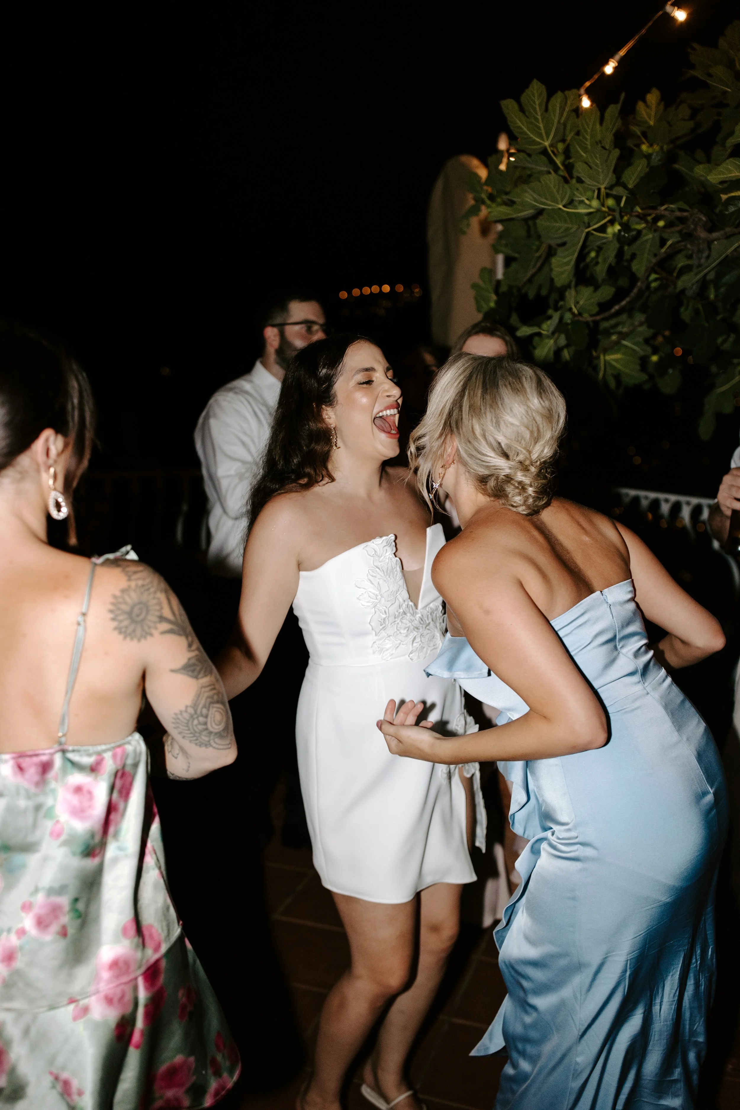 Group of women dancing and laughing at a nighttime outdoor event, with some wearing dresses and others casual attire, under string lights and greenery.