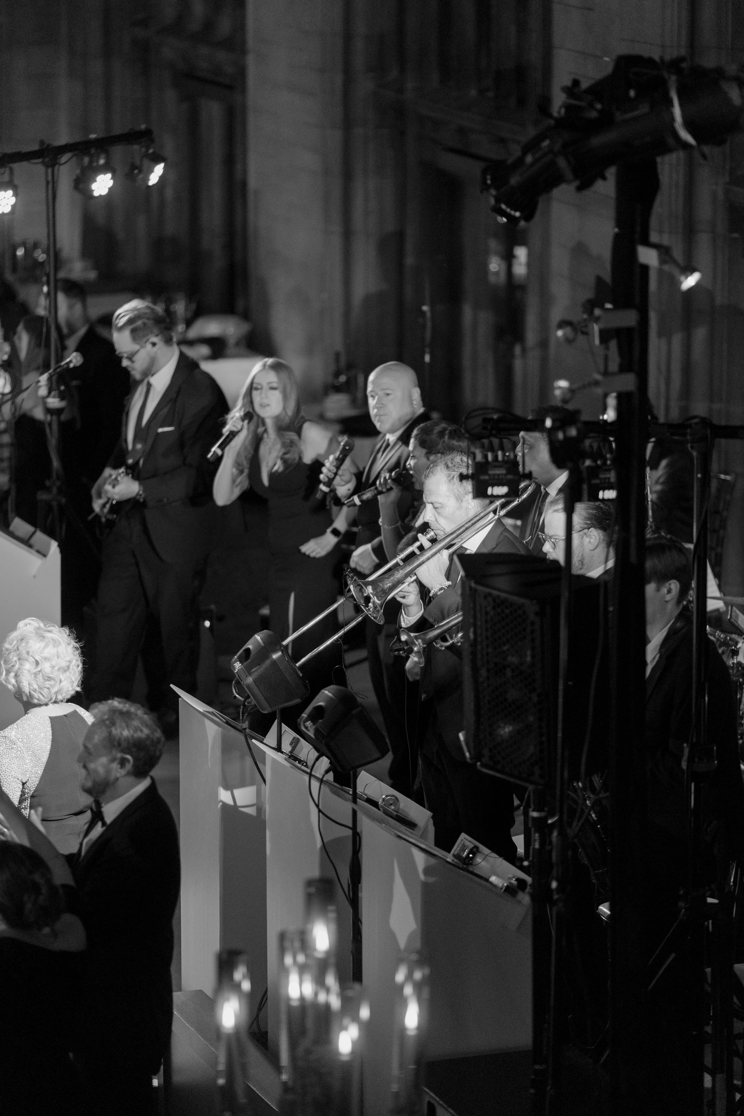 A black-and-white photo of a band performing on stage at an indoor venue. The band members are dressed in formal attire. Multiple people are seated in front of the stage, watching the performance.