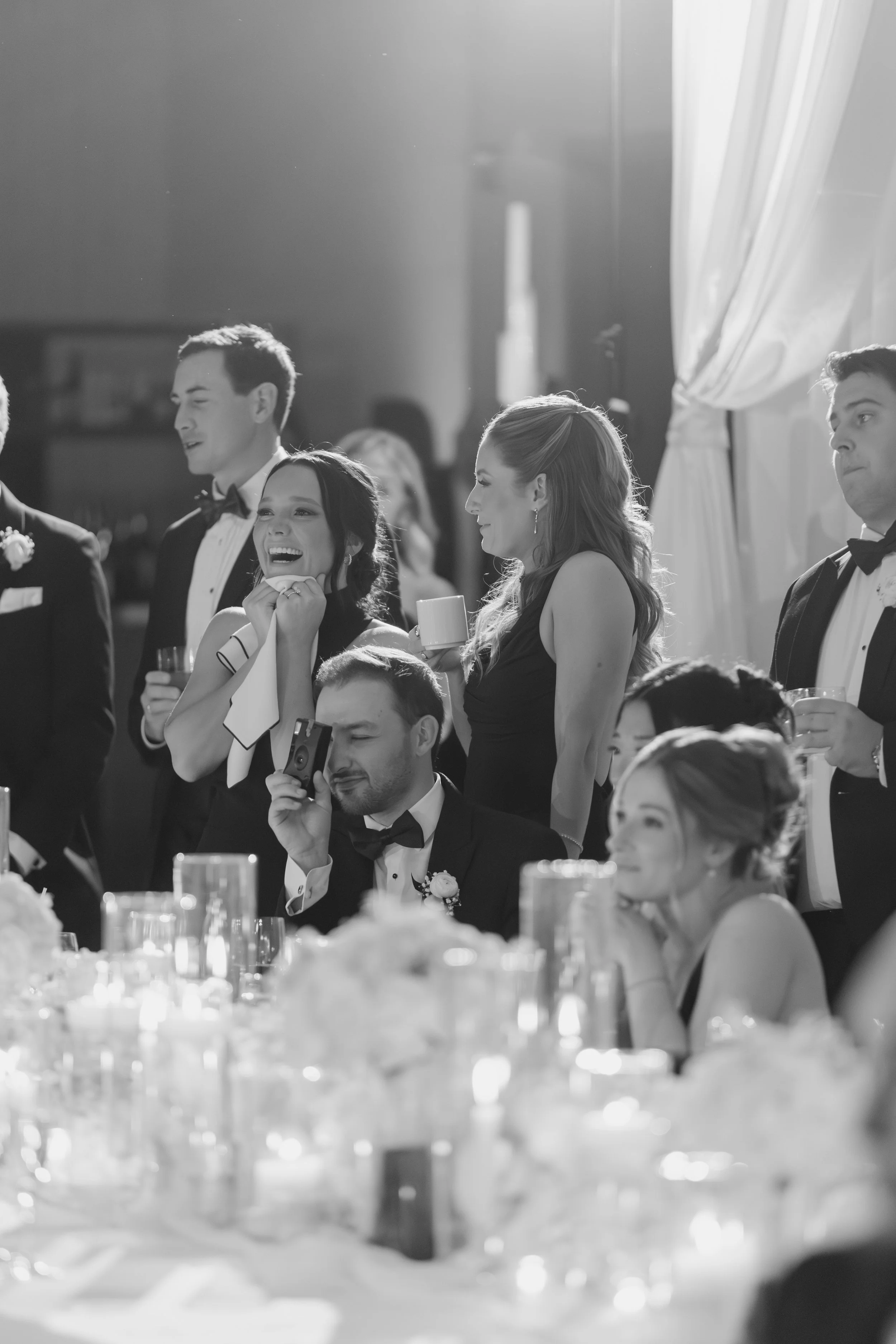 Black and white photo of people attending a formal event, seated at a table, and dressed in tuxedos and evening gowns. Some individuals are smiling, holding drinks, and engaging in conversations. The setting appears elegant with table decorations vis
