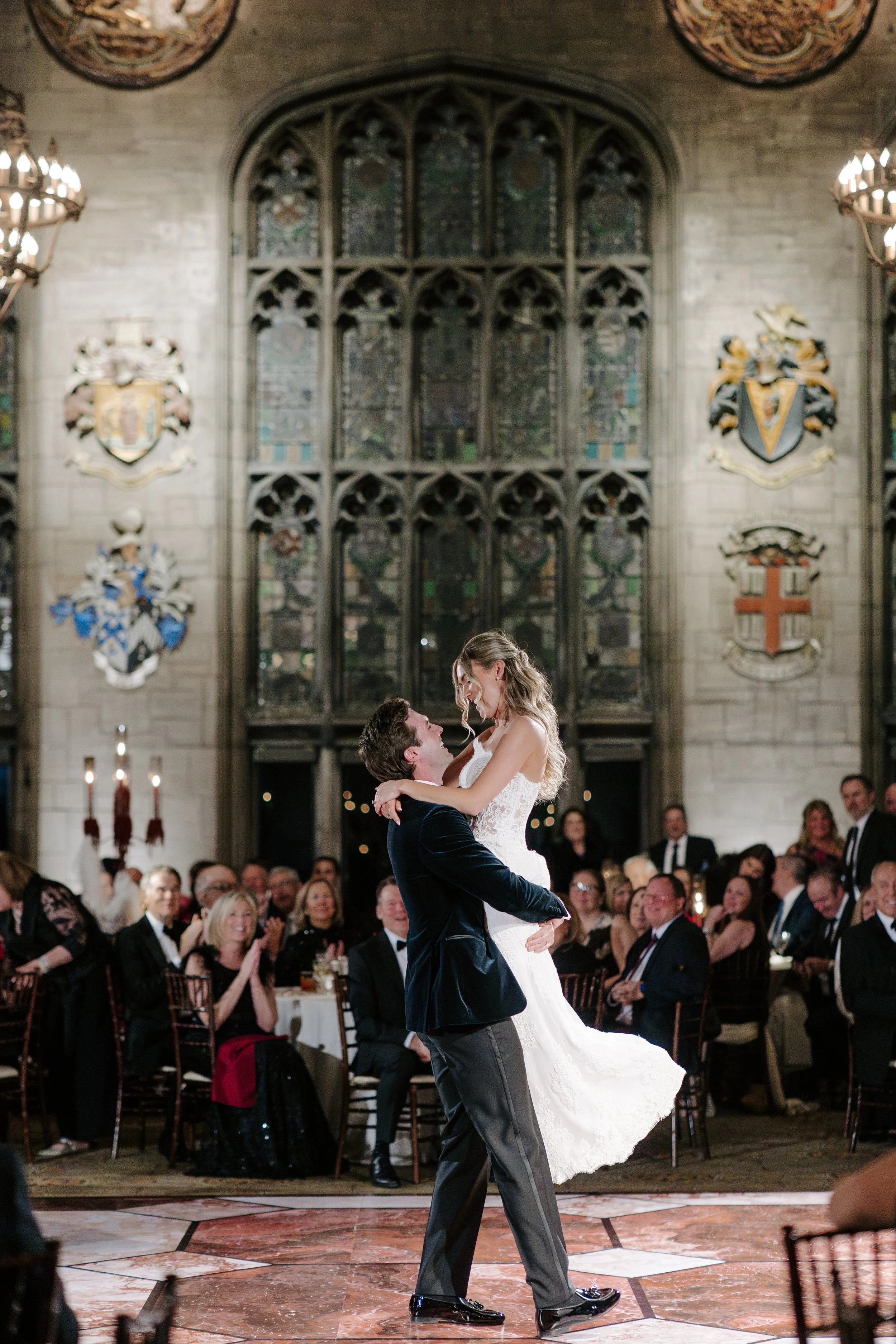 A couple dancing at their wedding reception in a grand hall with stained glass windows and coats of arms on the walls, surrounded by seated guests and live candles.