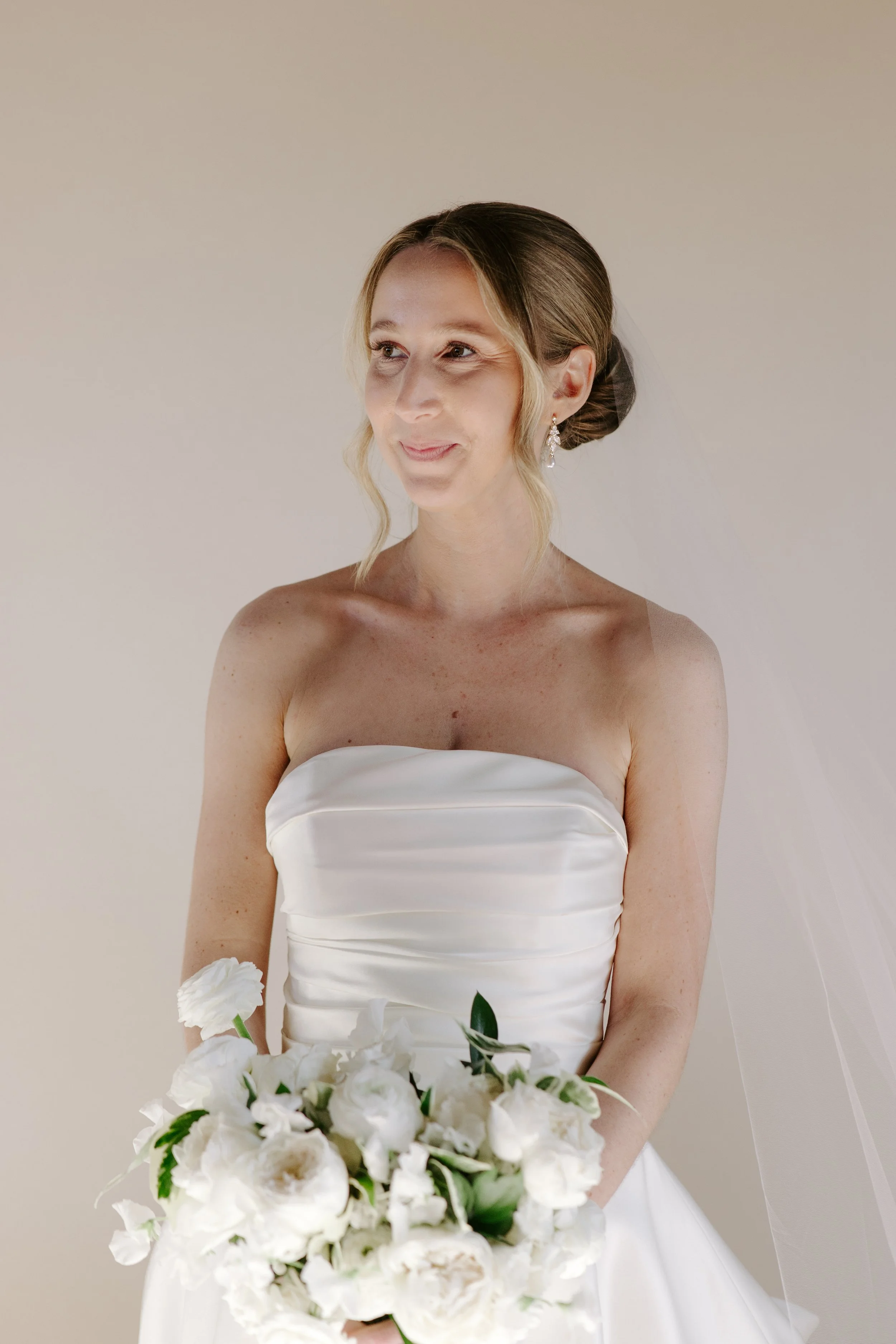 A woman in a strapless white wedding dress holding a bouquet of white flowers, with a black hair bun and drop earrings, standing against a plain light background.