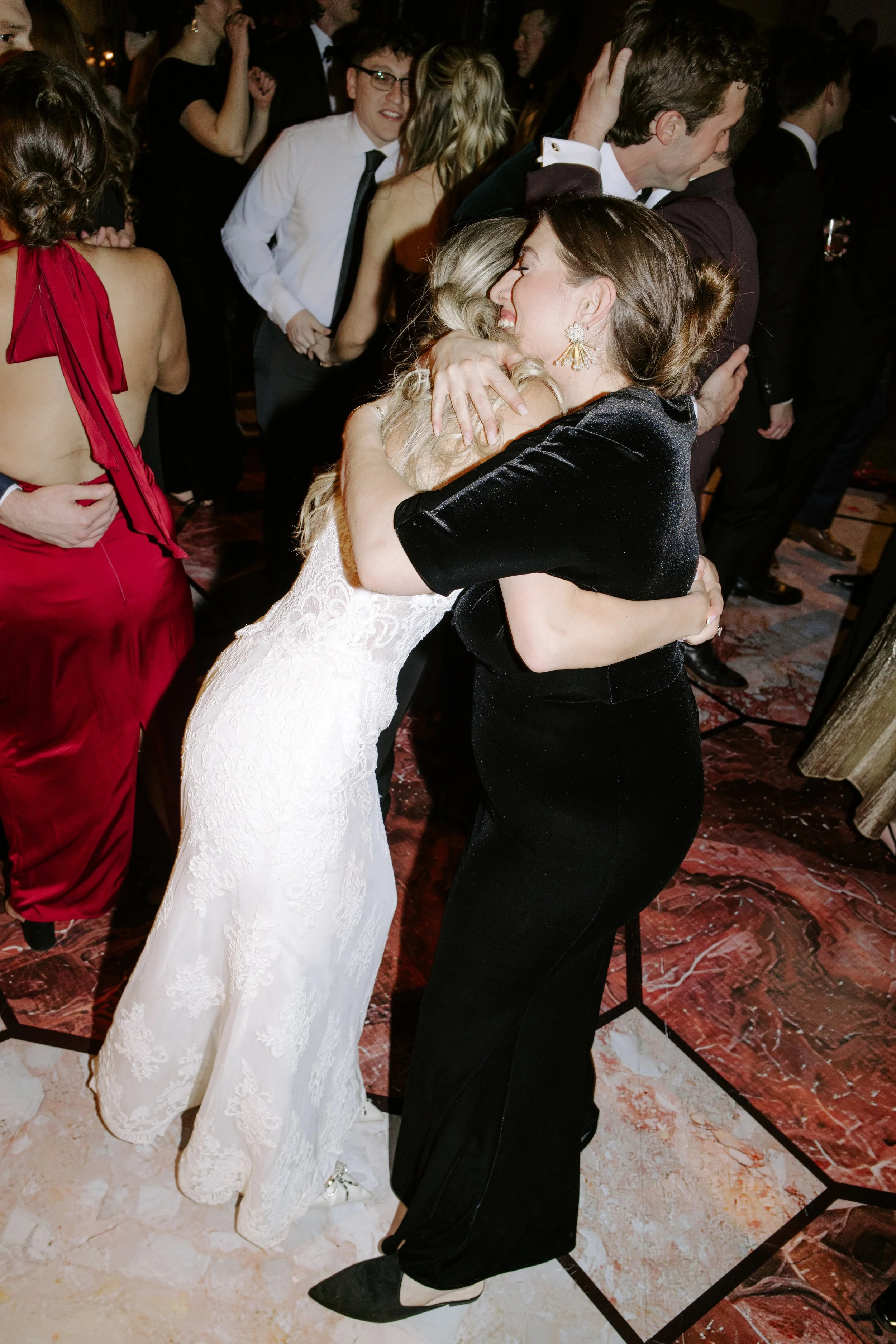 Two women hugging at a formal event, surrounded by people dancing and socializing.