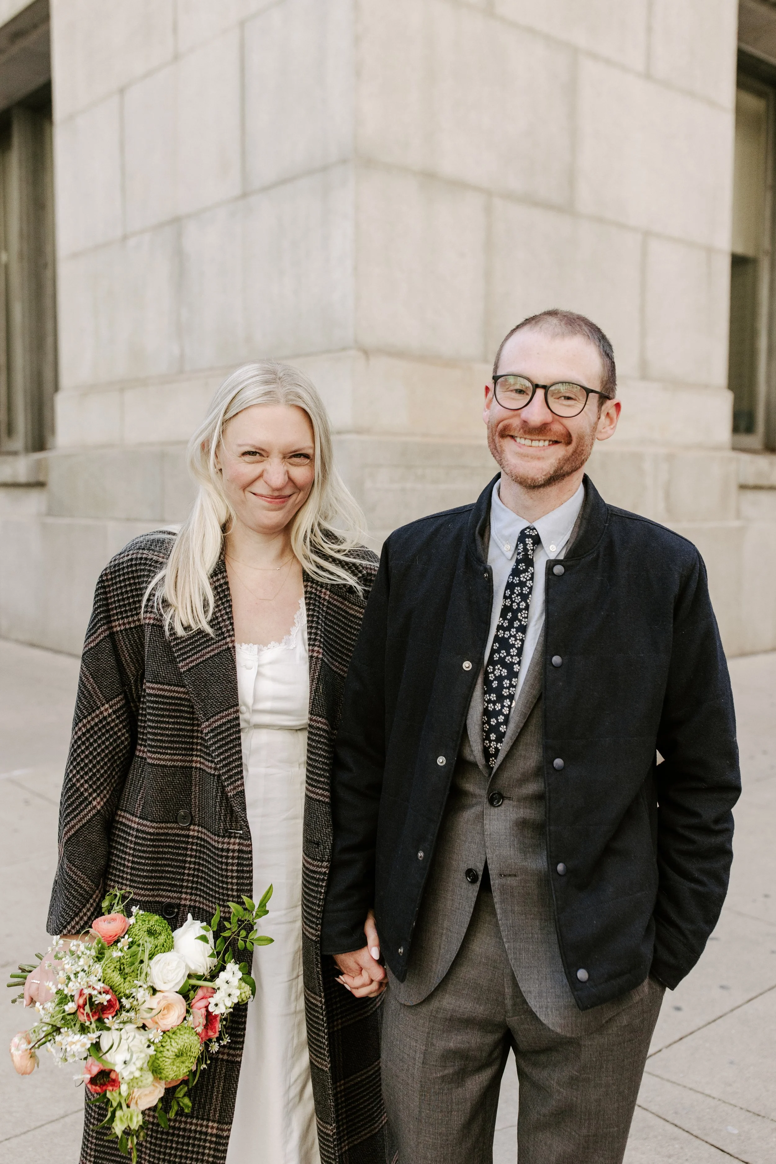 A smiling woman and a smiling man holding hands outside a building with beige stone walls. She is holding a bouquet of flowers.