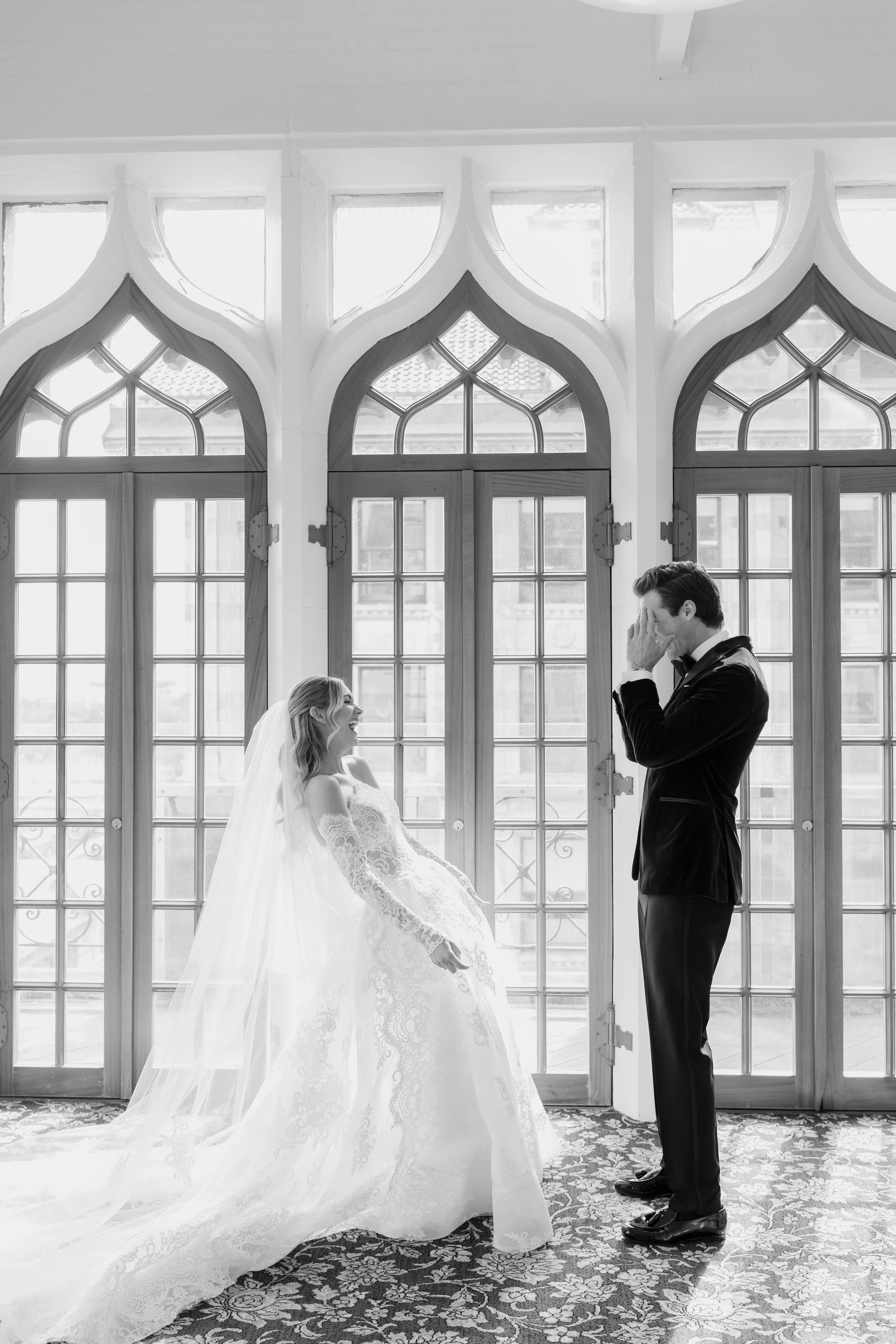 A bride and groom in wedding attire inside a room with large windows and decorative arches. The bride is seated, smiling, wearing a lace wedding gown and veil, while the groom is standing, smiling, with one hand covering his face in a gesture of emot