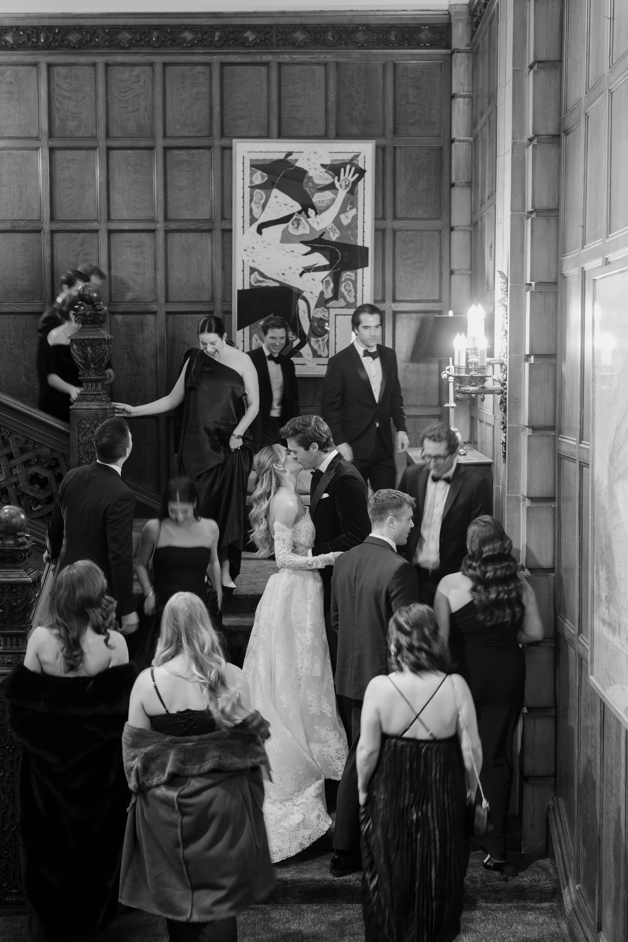 A formal black-and-white photograph of a wedding reception with guests ascending a staircase in an ornate, wood-paneled room. The bride and groom are near the center, engaging in a kiss, surrounded by well-dressed attendees in evening attire.