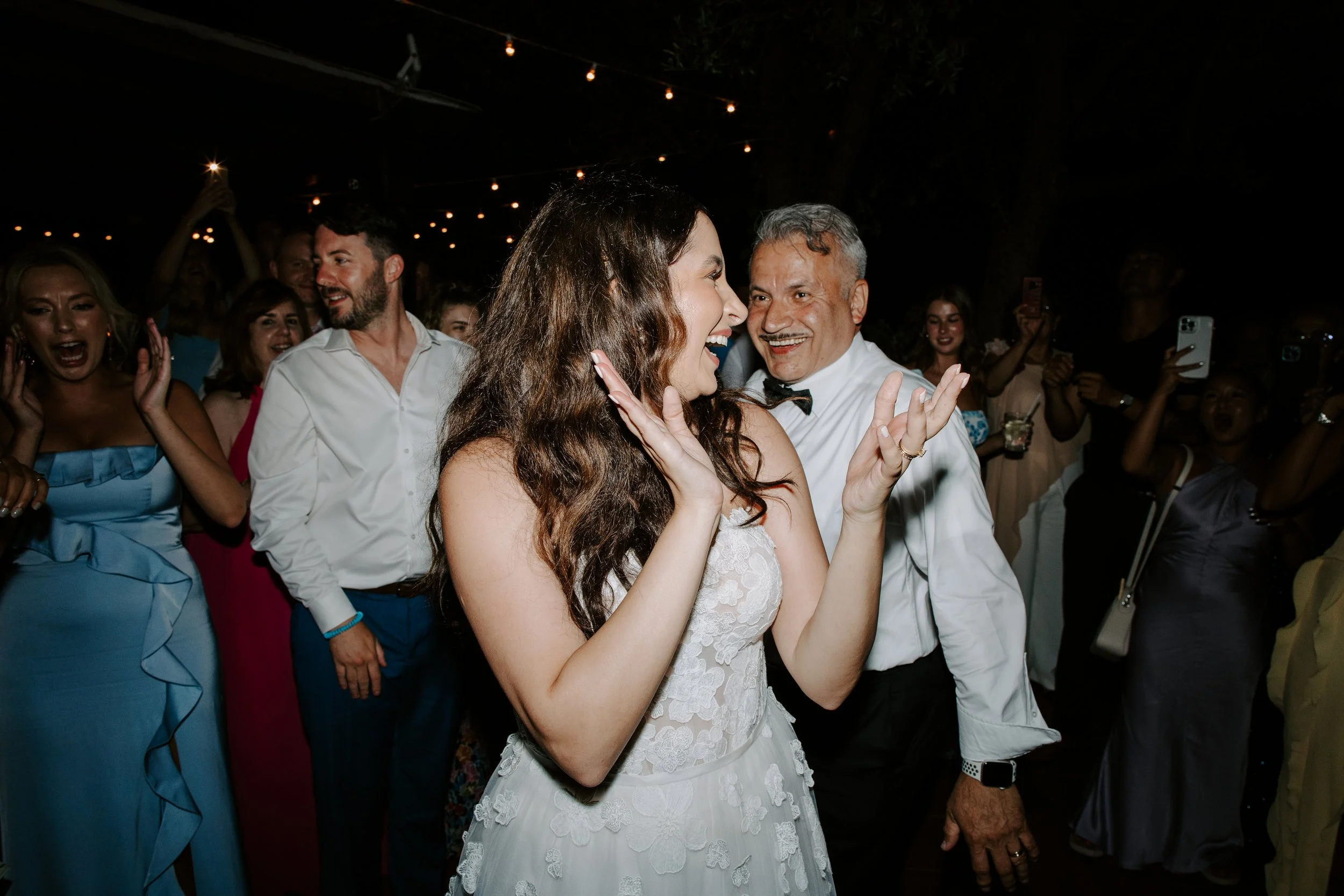 A group of people dressed in semi-formal attire celebrating at night under string lights. A woman in a white lace dress is smiling and laughing, sharing a joyful moment with an older man in a white shirt with a bow tie. Others in the background are a