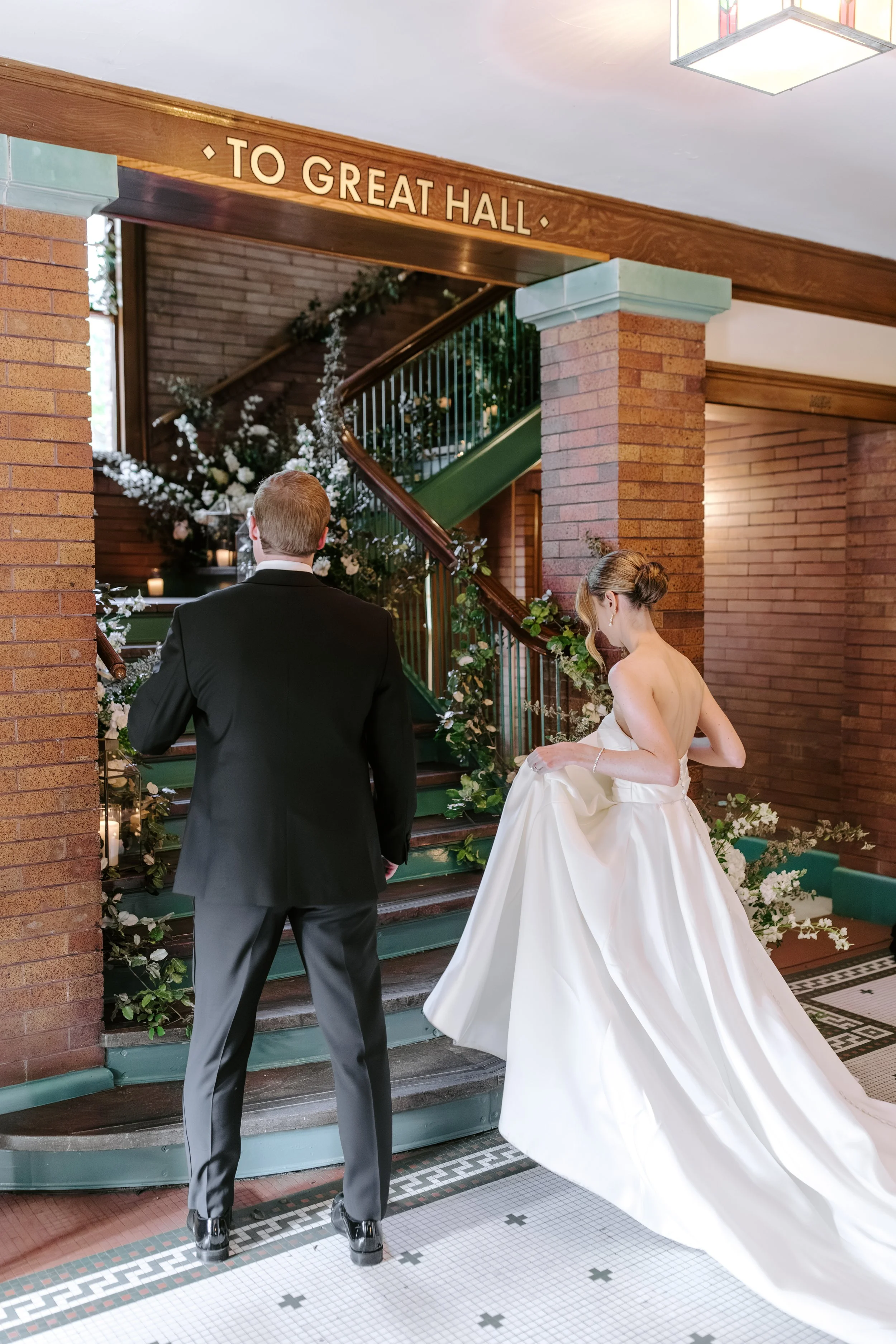 A bride and groom standing at the bottom of a staircase decorated with white flowers and candles, inside a brick building with a sign pointing to the great hall.