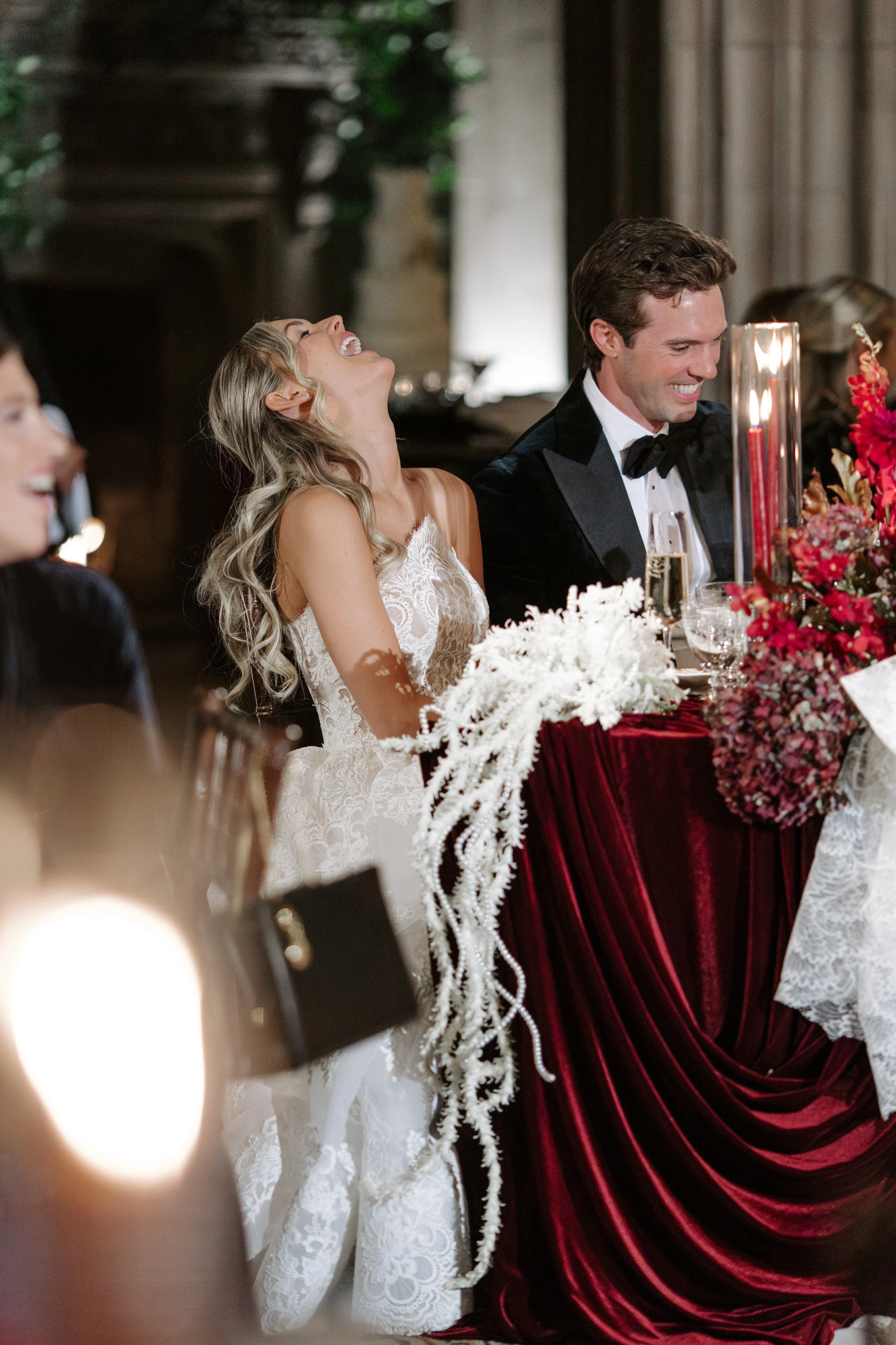 A joyful woman in a wedding dress laughing with her head tilted back while sitting next to a man in a tuxedo at a formal event, with flowers and candles on the table in front of them.