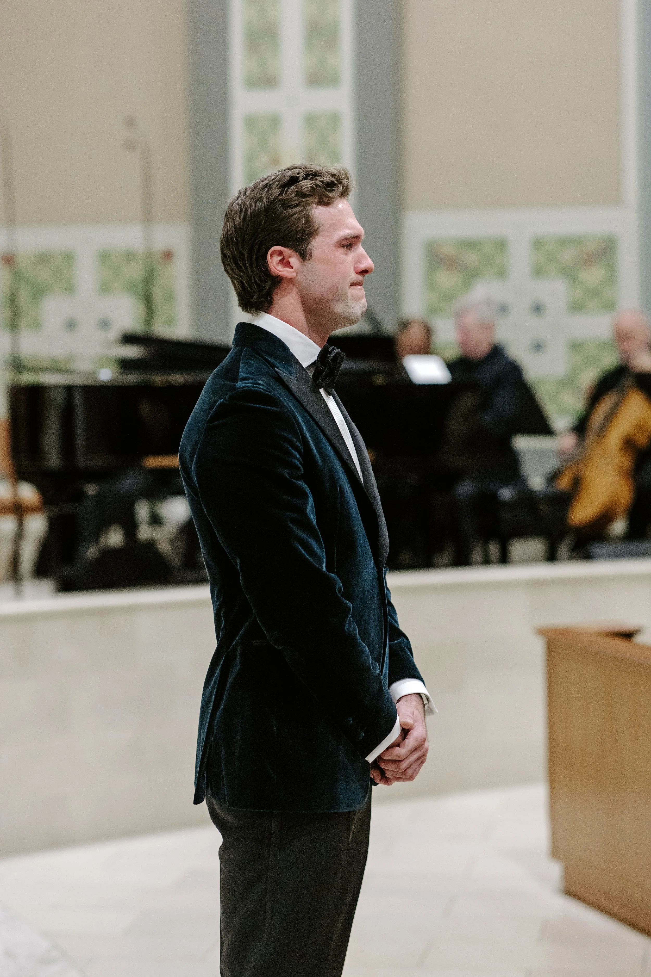 A man with brown hair dressed in a tuxedo standing in profile in a room with people and a piano in the background.
