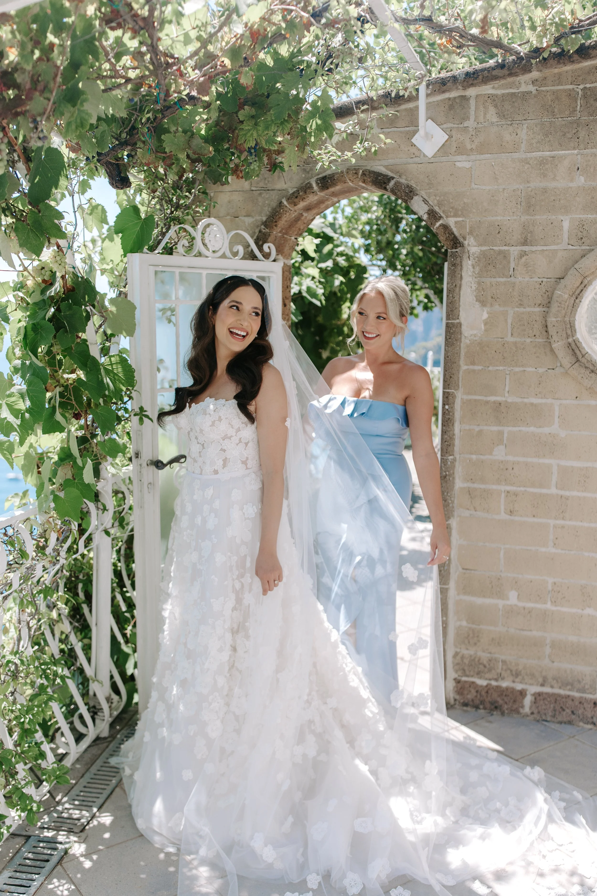 Two women, one in a white wedding gown and the other in a light blue dress, standing and smiling outdoors near a brick wall and an arched doorway, surrounded by greenery and flowers.