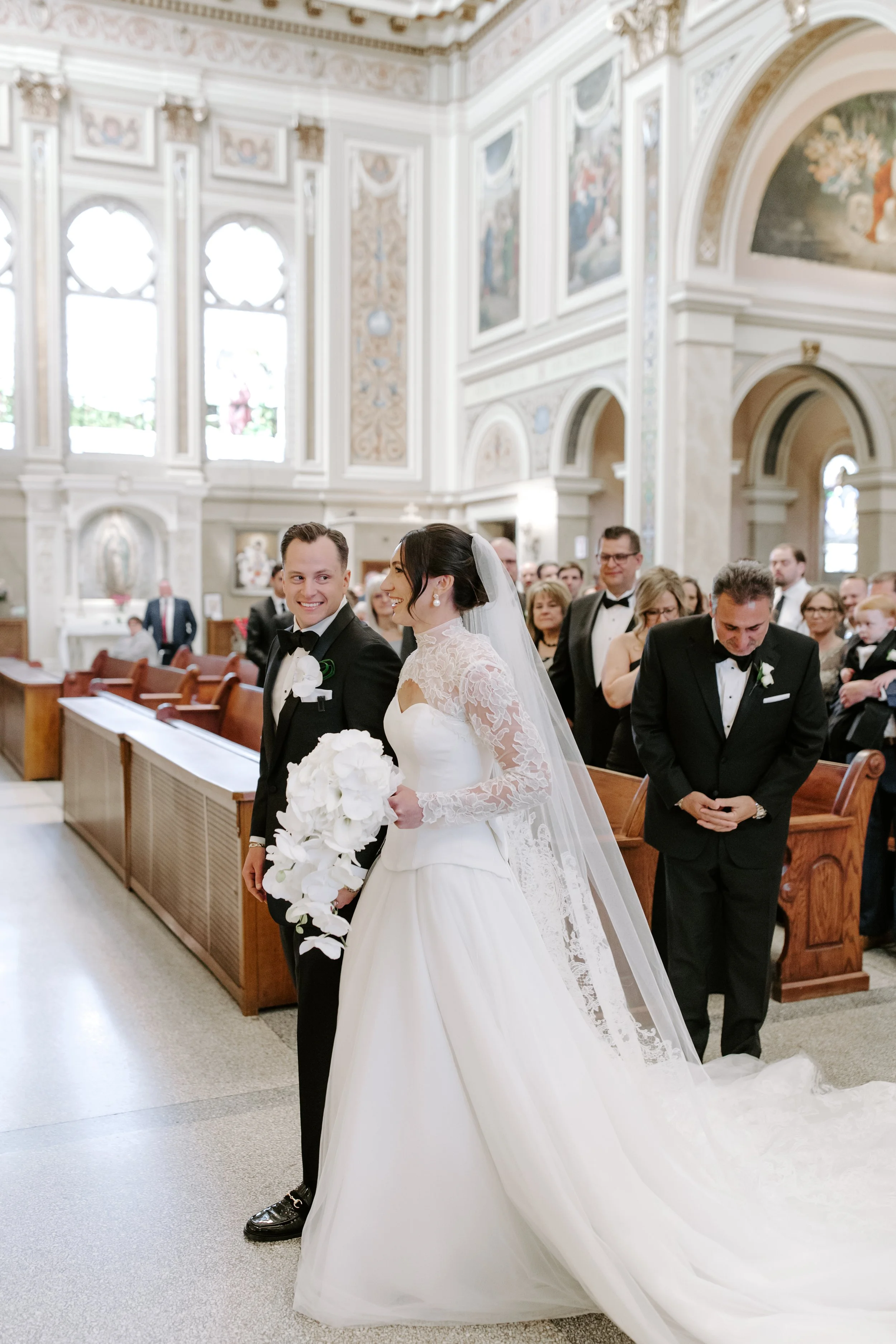 A bride and groom standing at the altar during a wedding ceremony inside a church, surrounded by guests, with high arched windows and ornate interior decor.