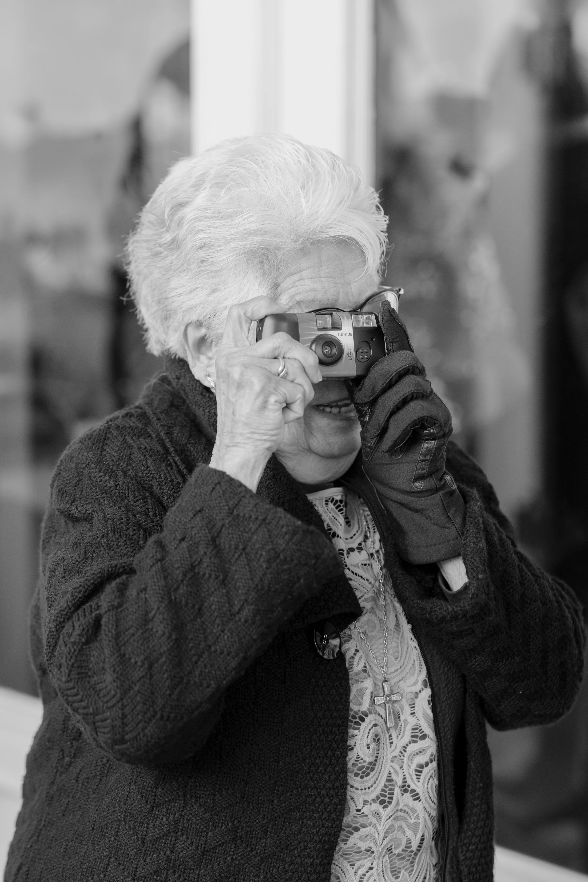 An elderly woman with white hair taking a photo with a small camera, wearing glasses and a patterned blouse under a dark cardigan, standing indoors.