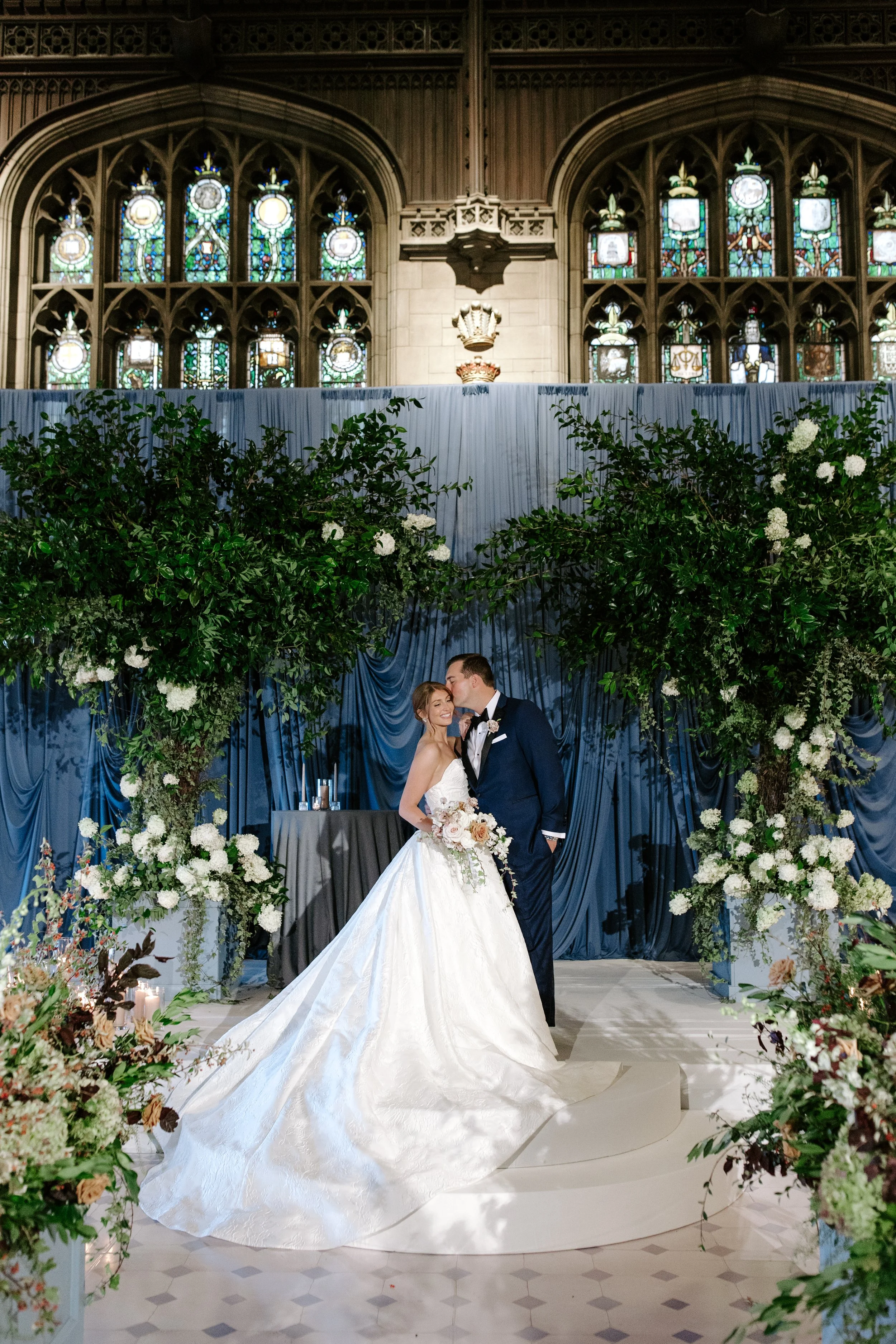 A bride and groom kissing during their wedding ceremony inside a historic church with stained glass windows, decorated with greenery and white flowers.