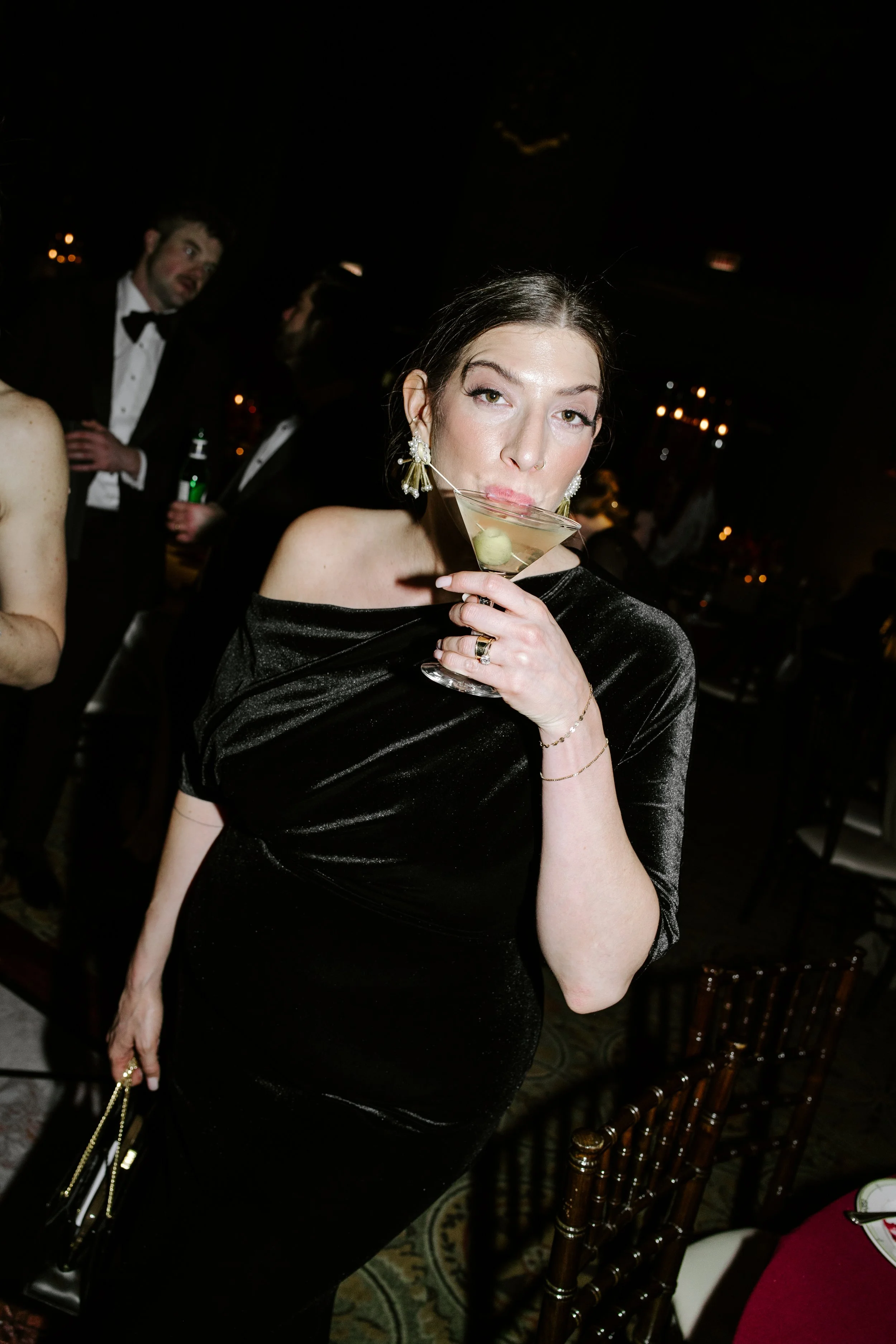 A woman in a black off-shoulder dress holding a martini glass at a formal event with other guests in tuxedos in the background.