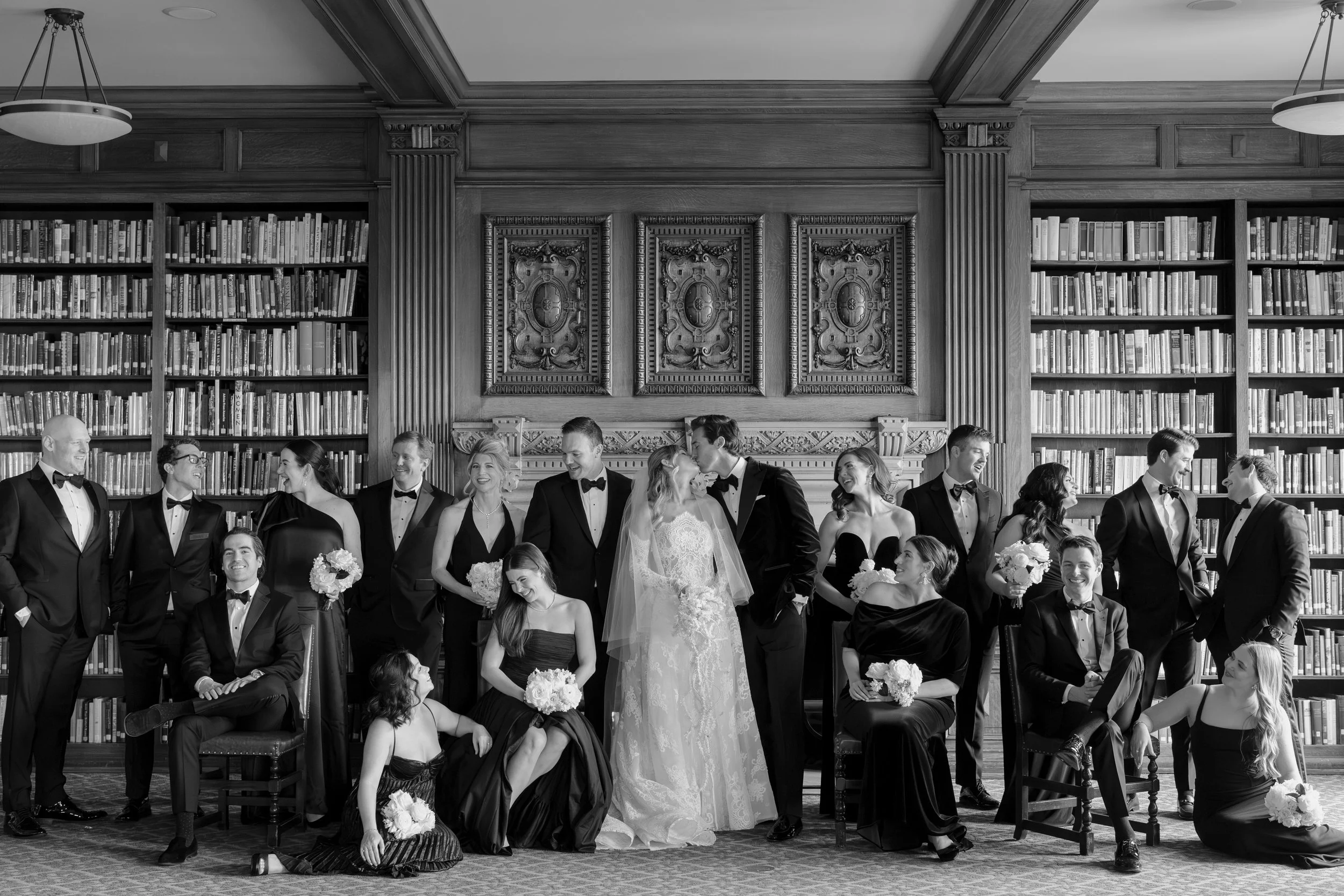 A group of wedding party members dressed in formal attire, standing and sitting in a grand library with high bookshelves and ornate woodwork, during a wedding photoshoot.