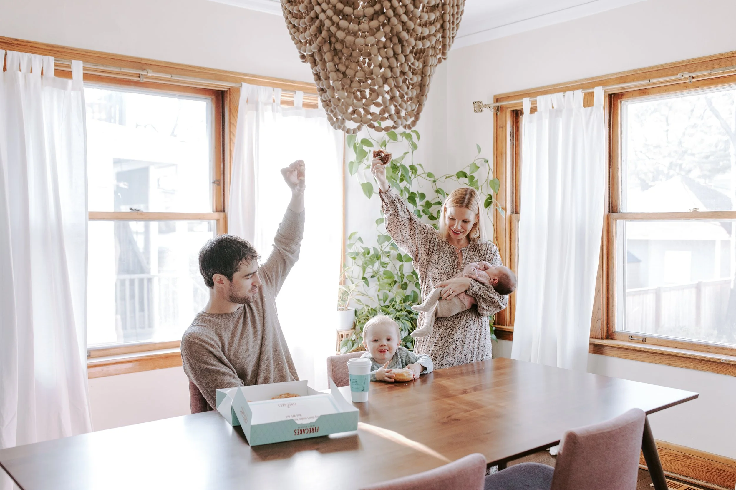 A family gathering around a dining table in a bright room with large windows and white curtains. A woman holds a baby, while a man sits at the table with a young child. The table has food and a pizza box.