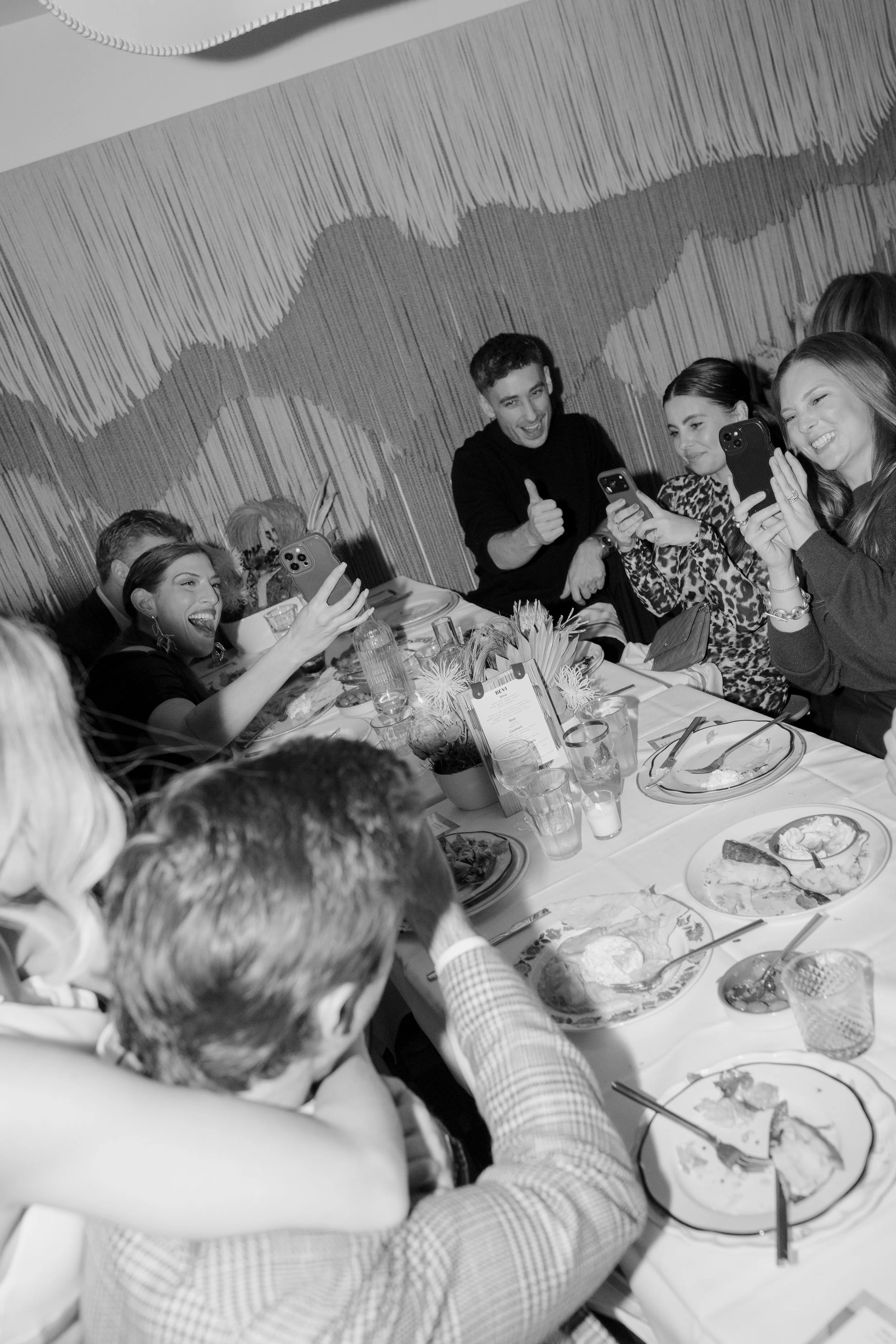 Group of people sitting around a table taking photos with smartphones, enjoying a meal, in a restaurant with a textured wall pattern.