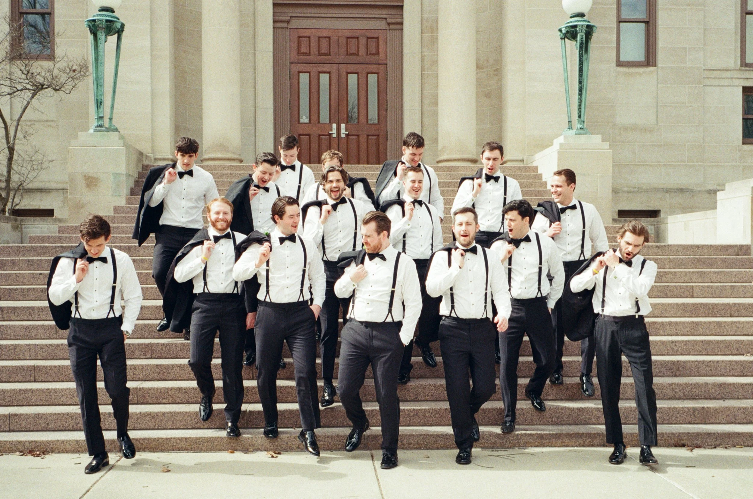A group of young men dressed in tuxedos and carrying black bags walking down steps in front of a building with large wooden double doors.