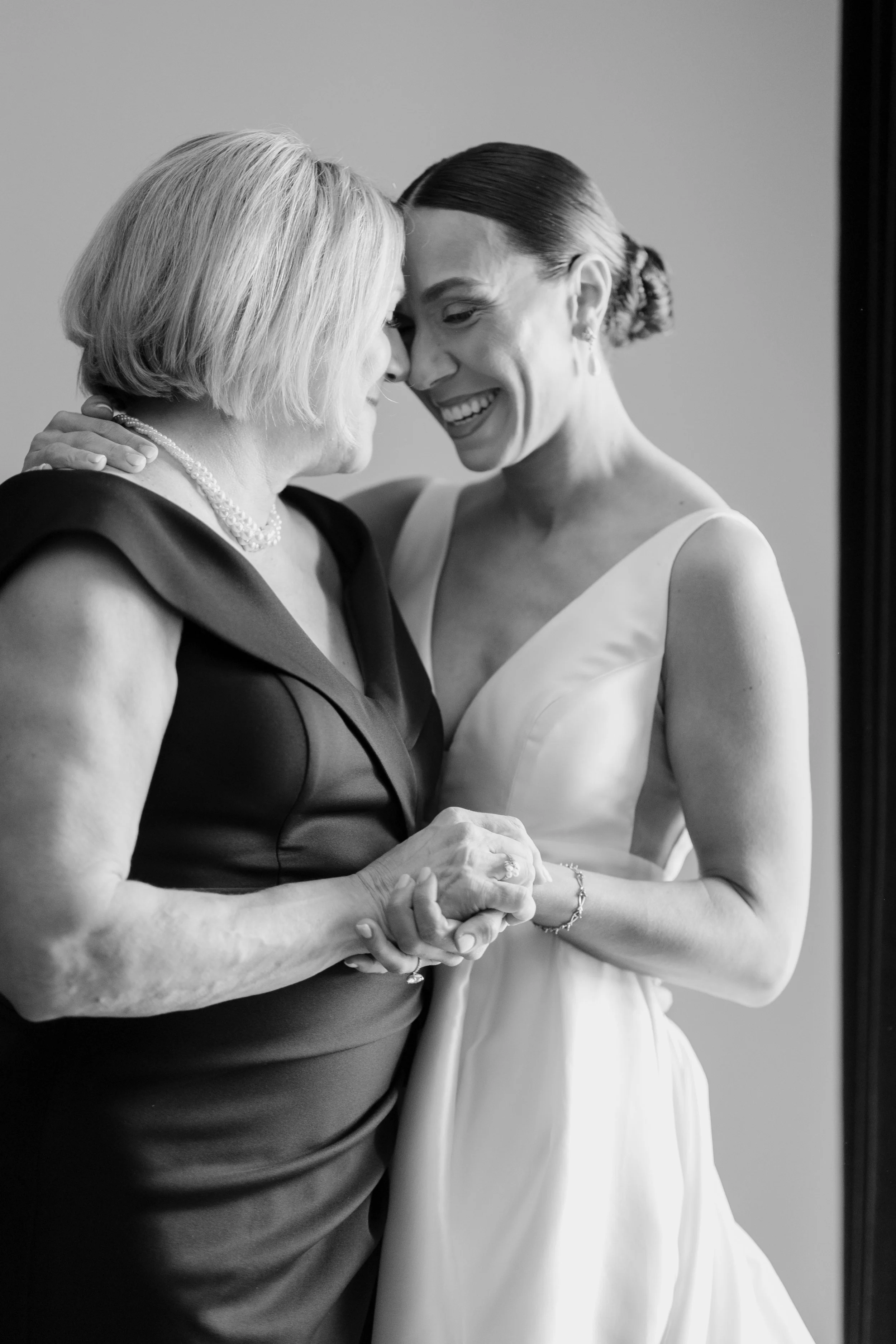 A Black-and-white photograph of two women embracing and smiling at each other. One woman, with blonde hair, is wearing a dark dress and a pearl necklace, while the other woman, with dark hair styled in a bun, is wearing a light-colored dress. They ar