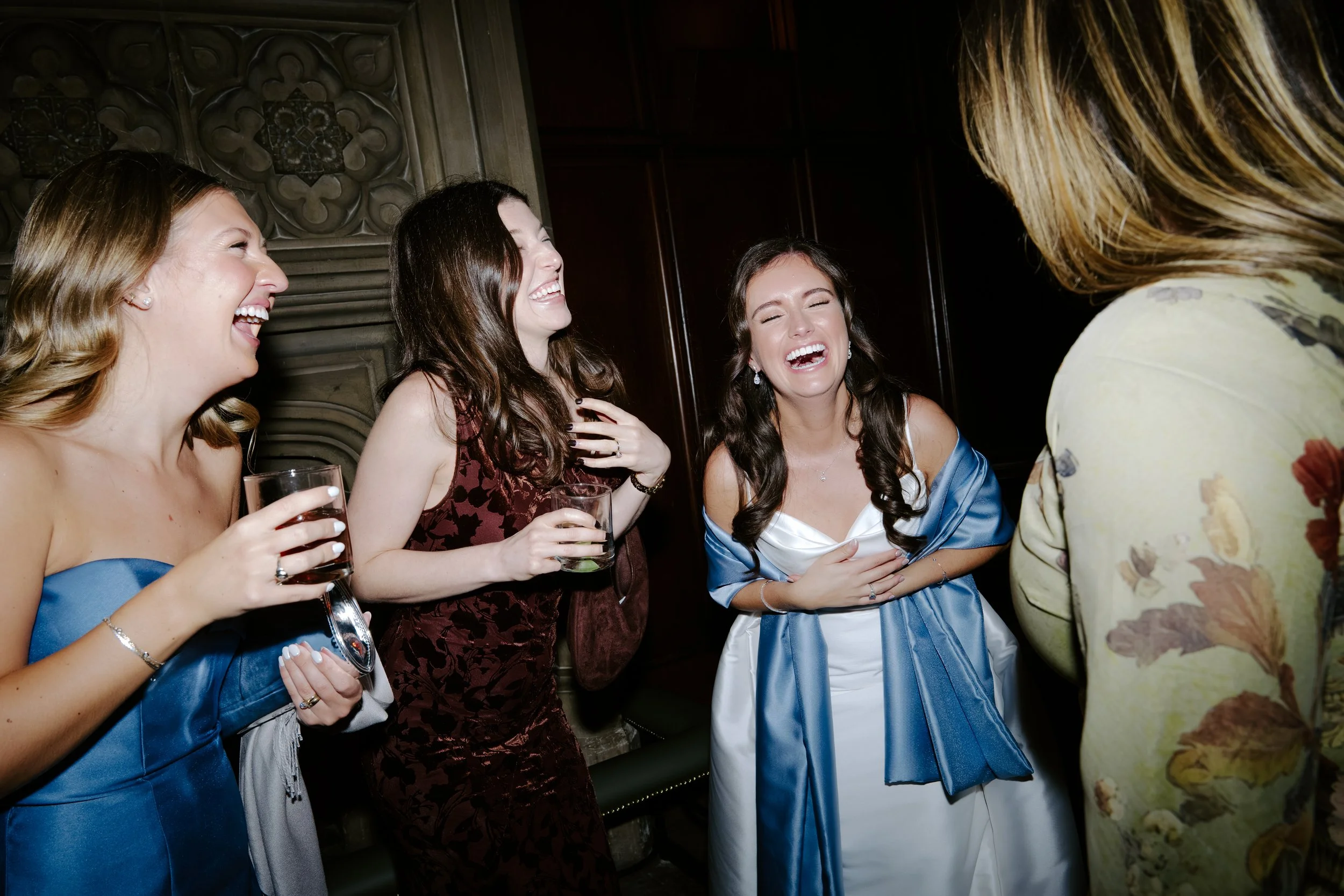 Group of women laughing and socializing at a party, holding glasses of drinks, with one woman wearing a blue satin dress and another in a floral dress.