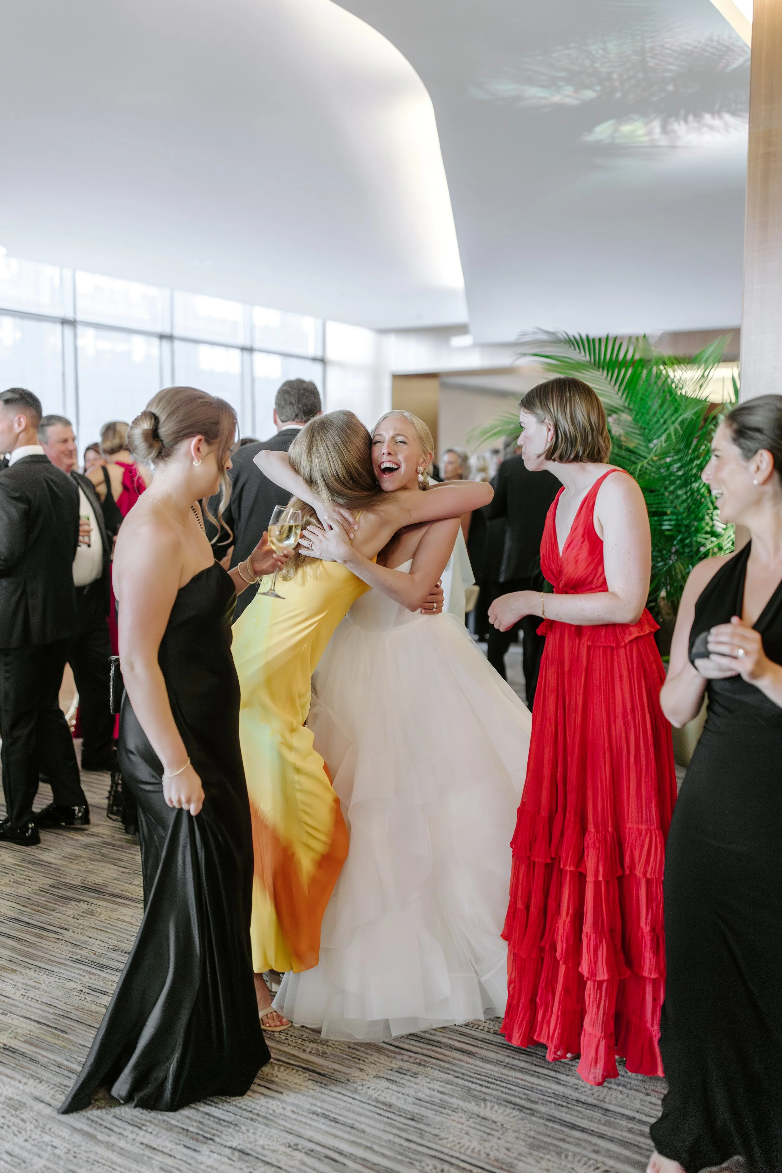 People at a social event, some hugging and holding glasses of champagne, in a bright and modern indoor space with large windows and greenery.