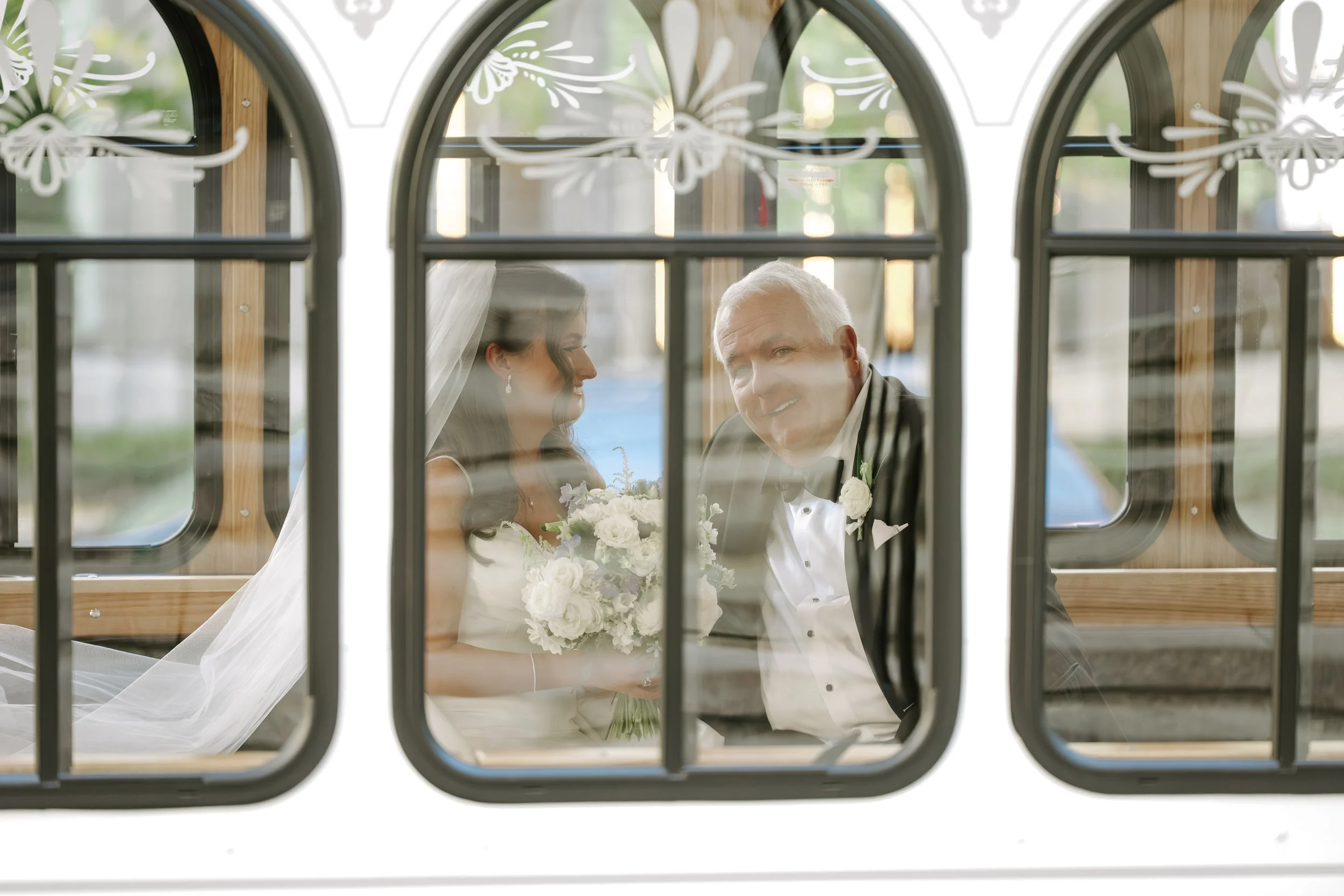 A bride and groom sitting inside a wooden church, viewed through decorative window panes with floral motifs, sharing a moment during their wedding.