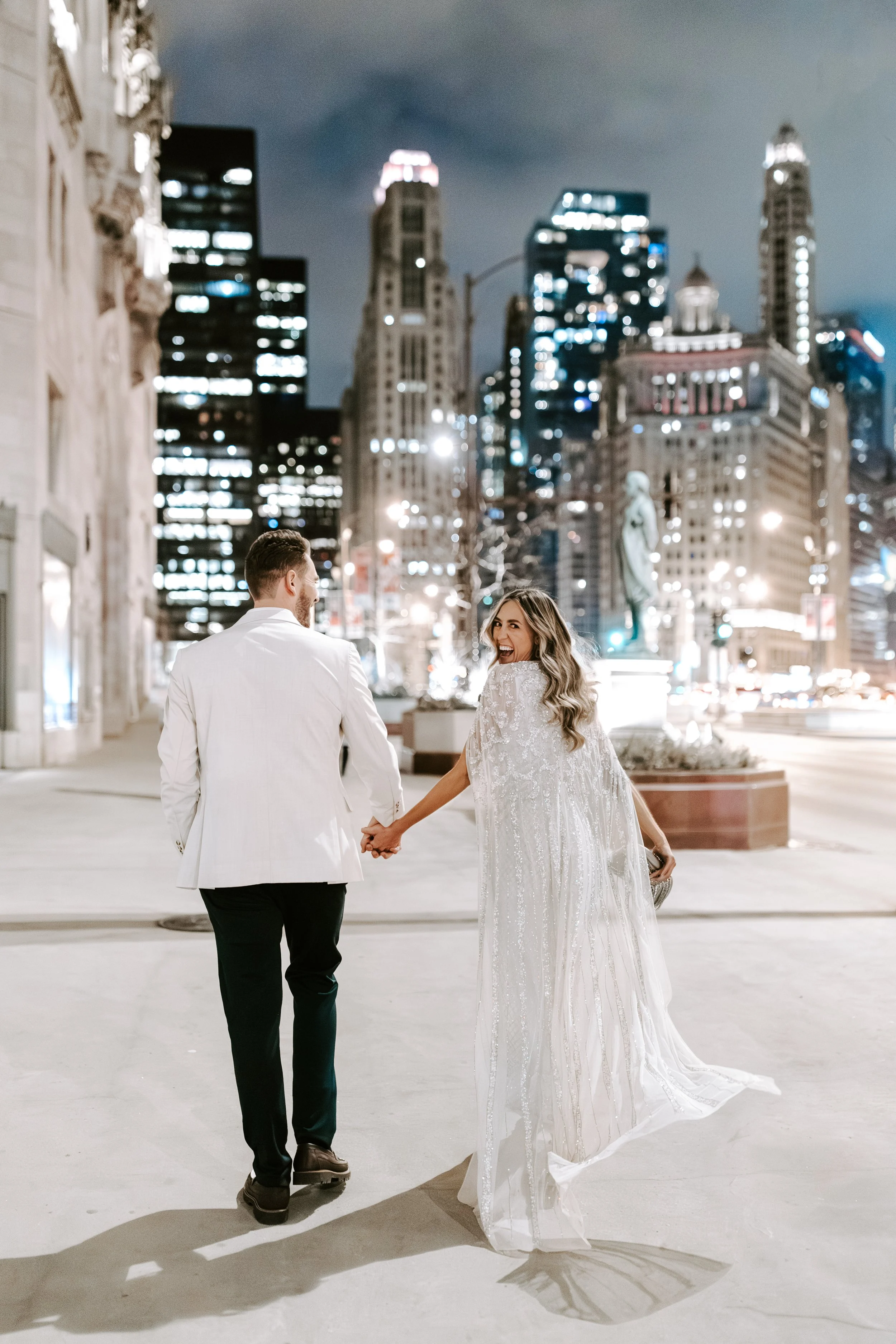 A newlywed couple walking hand in hand at night in a city with tall buildings lit up behind them. The woman is in a white wedding dress and the man is in a white suit jacket and black pants.