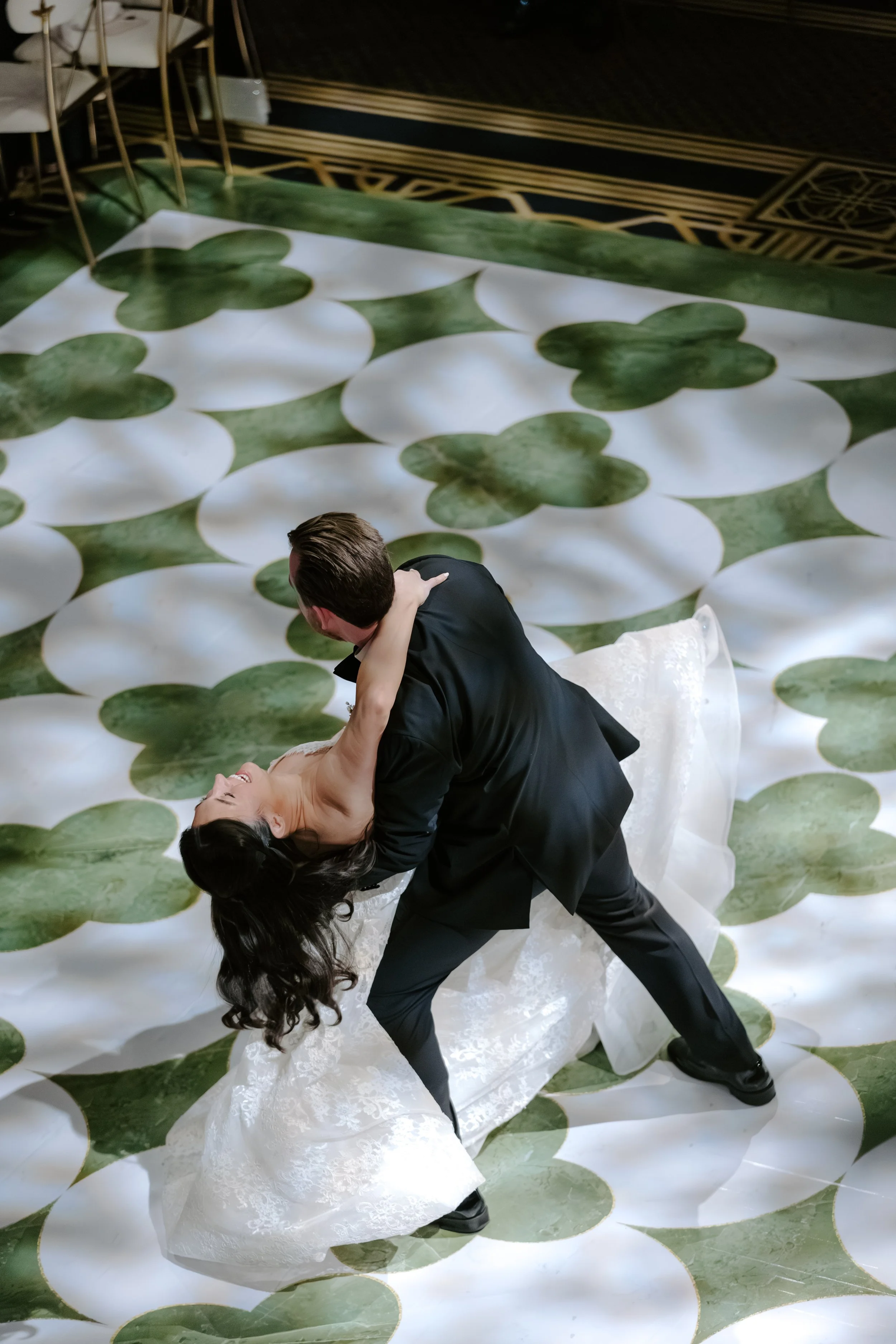 A couple dances in wedding attire on a patterned green and white floor, with the groom dipping the bride while holding her waist and the bride leaning back with her eyes closed and hair down.