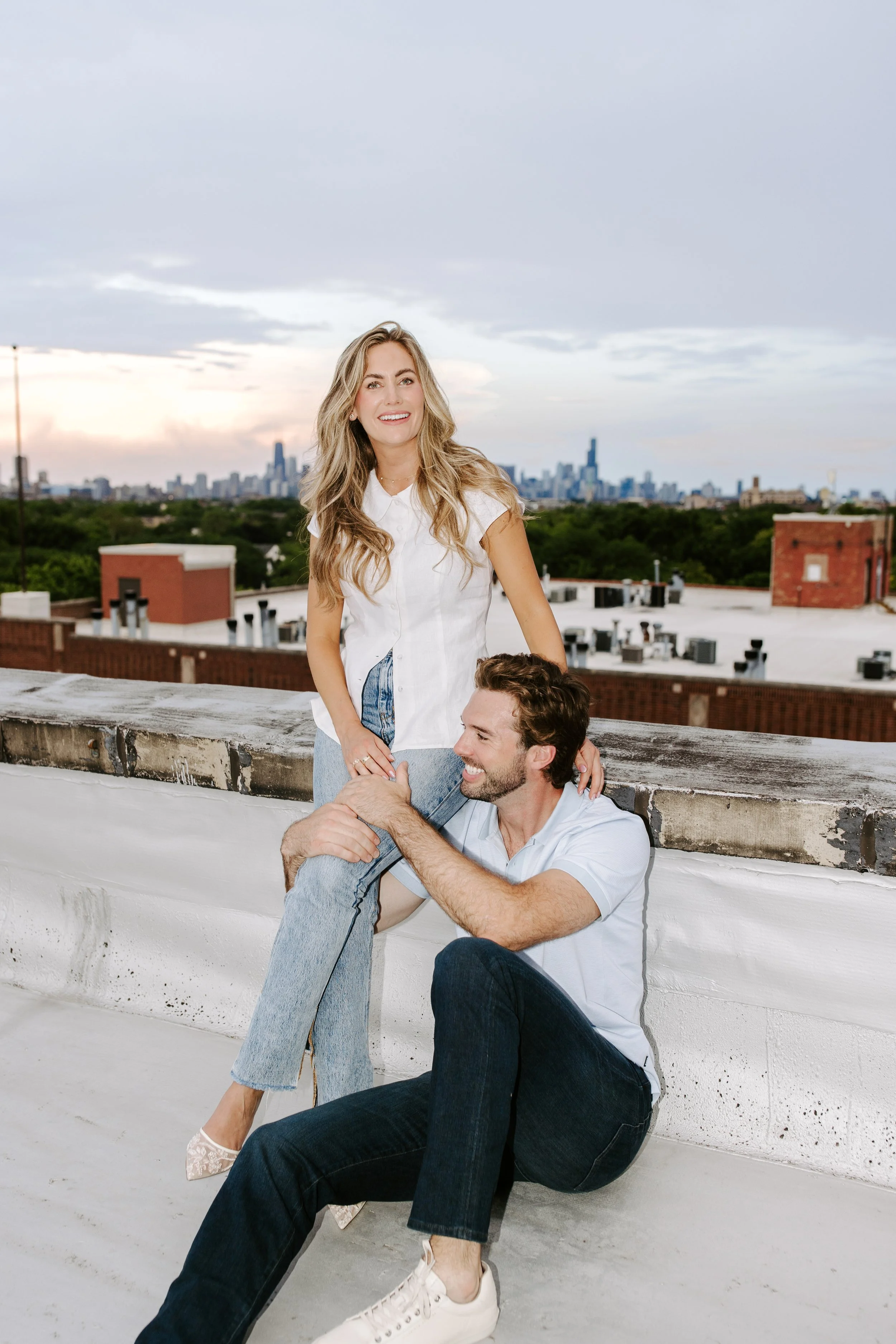 A young woman with blonde hair wearing a white shirt and jeans standing on a rooftop in front of a city skyline at sunset, smiling at the camera. A young man with brown hair and a beard sitting on a ledge, wearing a white shirt and dark jeans, lookin