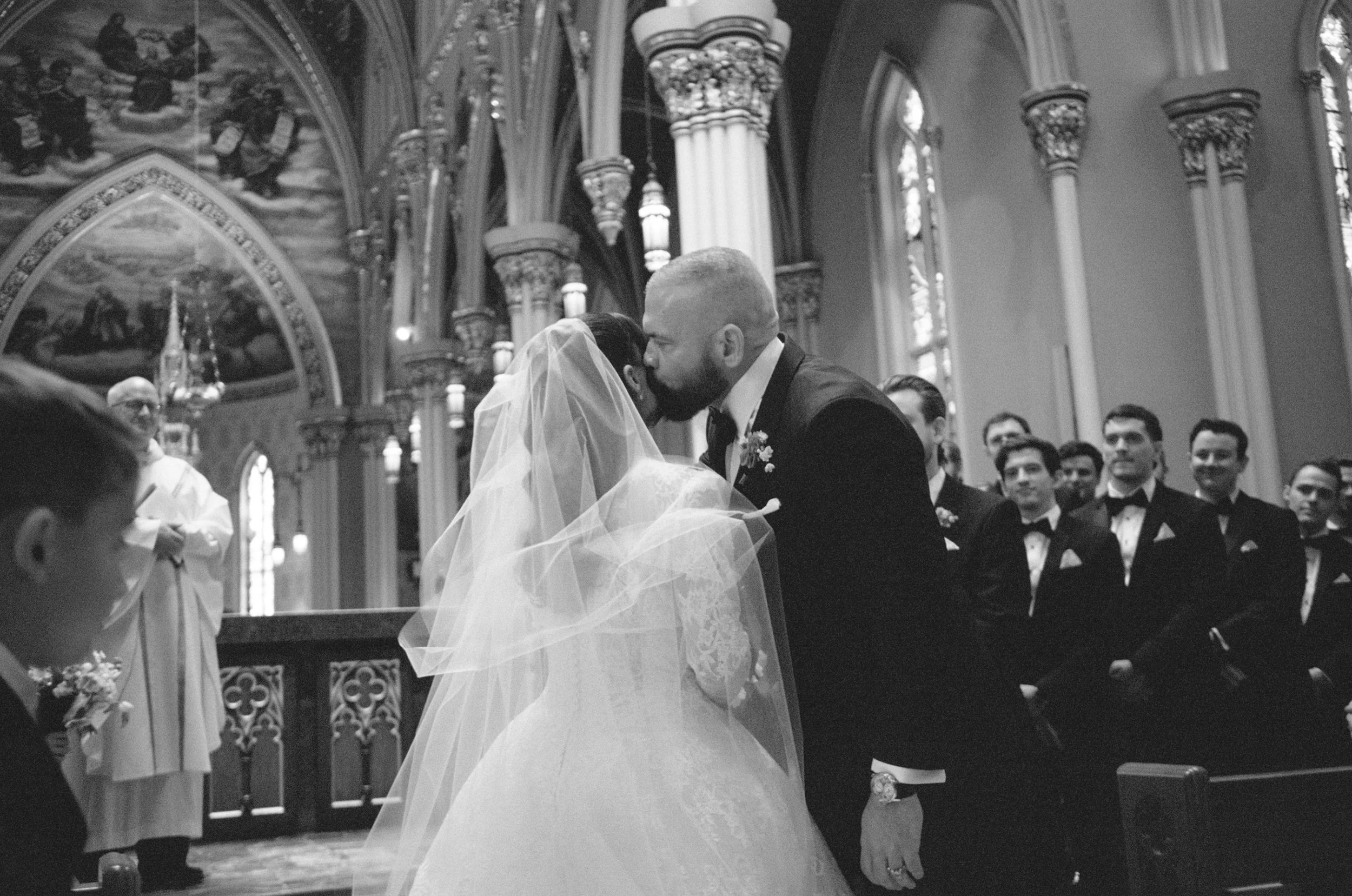 A bride and groom are sharing a kiss during their wedding ceremony in a church with ornate stained glass windows and columns, while guests watch.