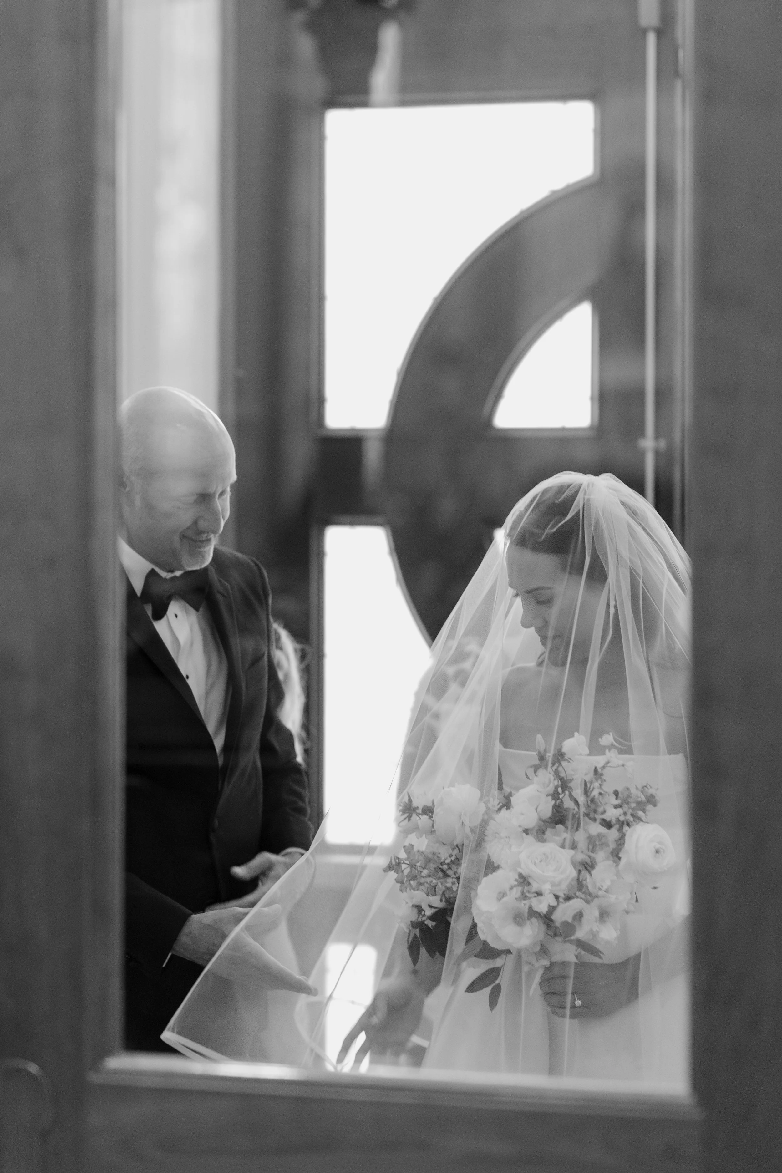 Black and white photo of a bride and groom during their wedding ceremony, standing inside a church, holding hands and facing each other, with the groom smiling and the bride holding a bouquet of flowers.