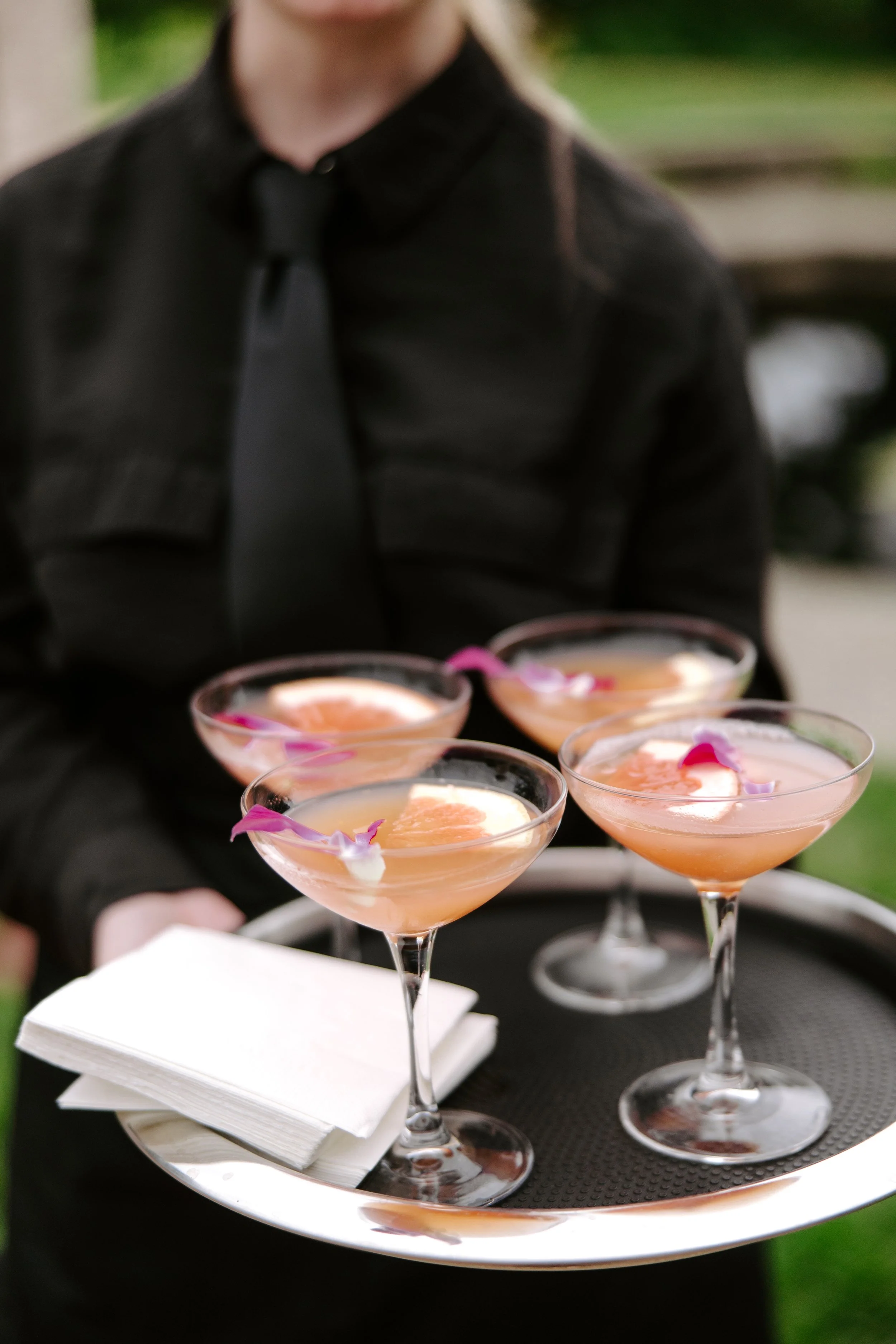 A person in a black shirt and tie holding a round tray with several pink cocktails garnished with purple flower petals, with a napkin underneath the tray.