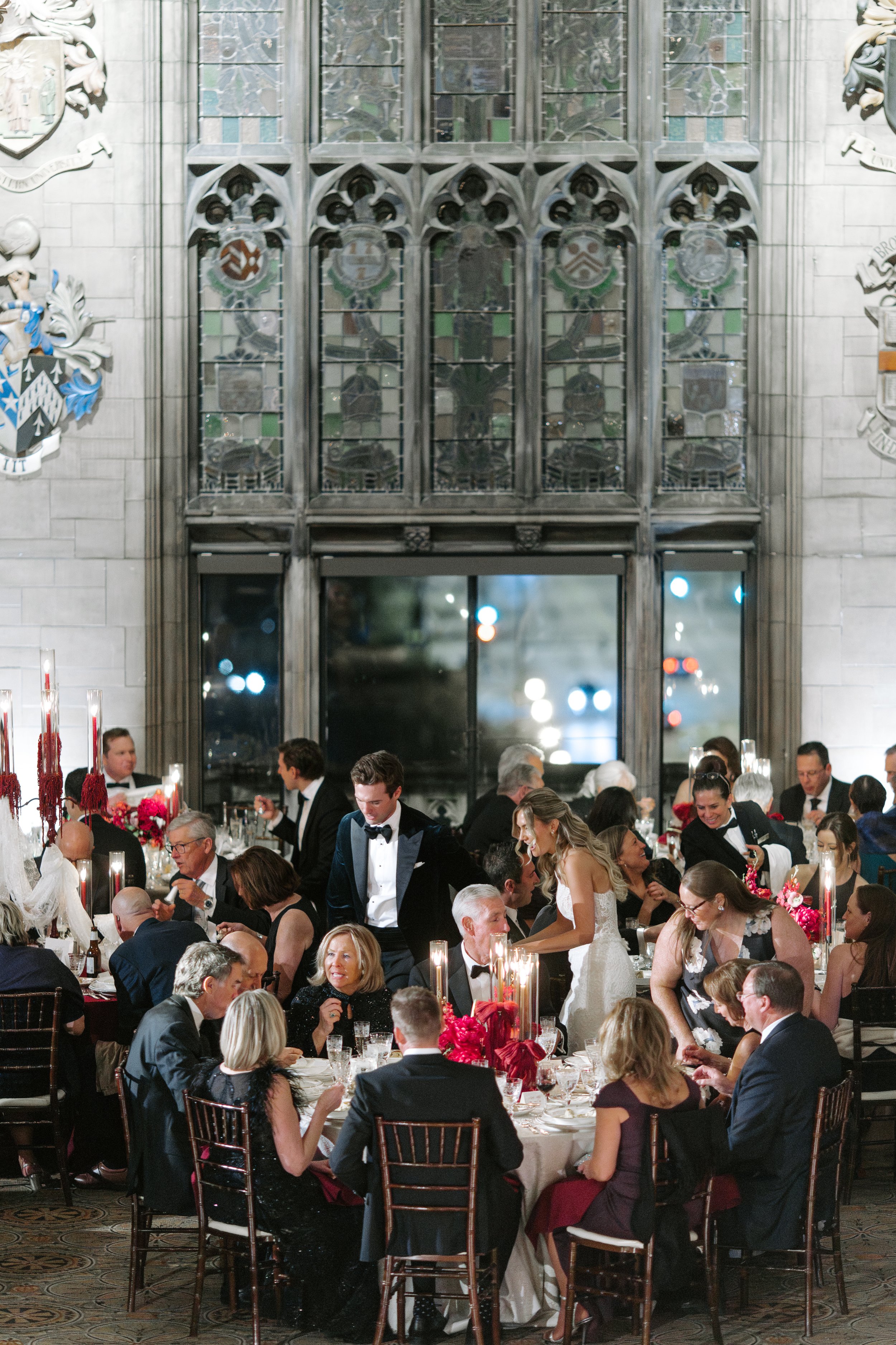 A wedding reception with guests seated at decorated tables, some standing and serving, in a grand hall with stained glass windows and heraldic shields on the walls.