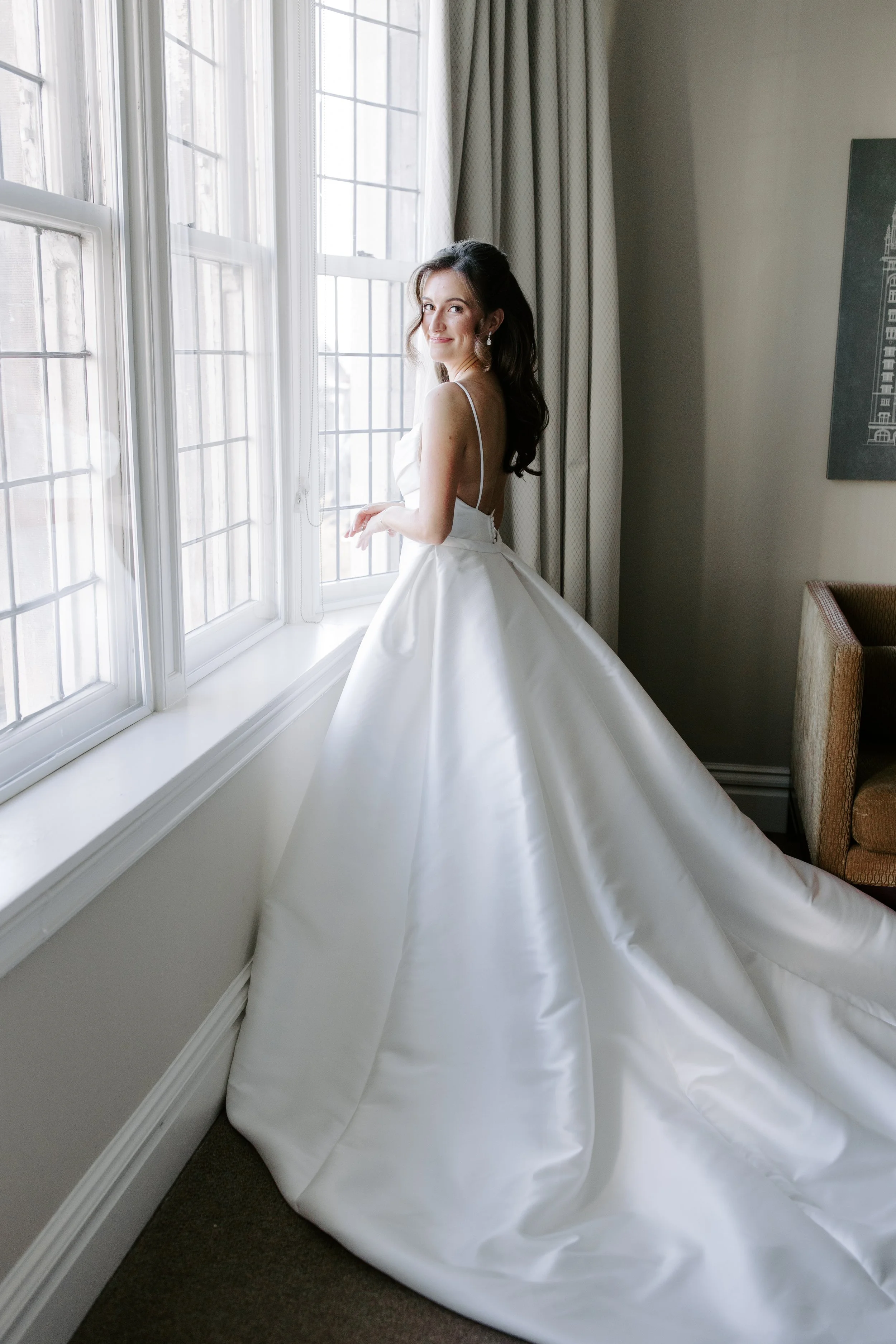 A woman in a white wedding dress is standing by a window with sheer curtains, smiling and looking over her shoulder inside a room with neutral-colored walls and a dark carpeted floor.