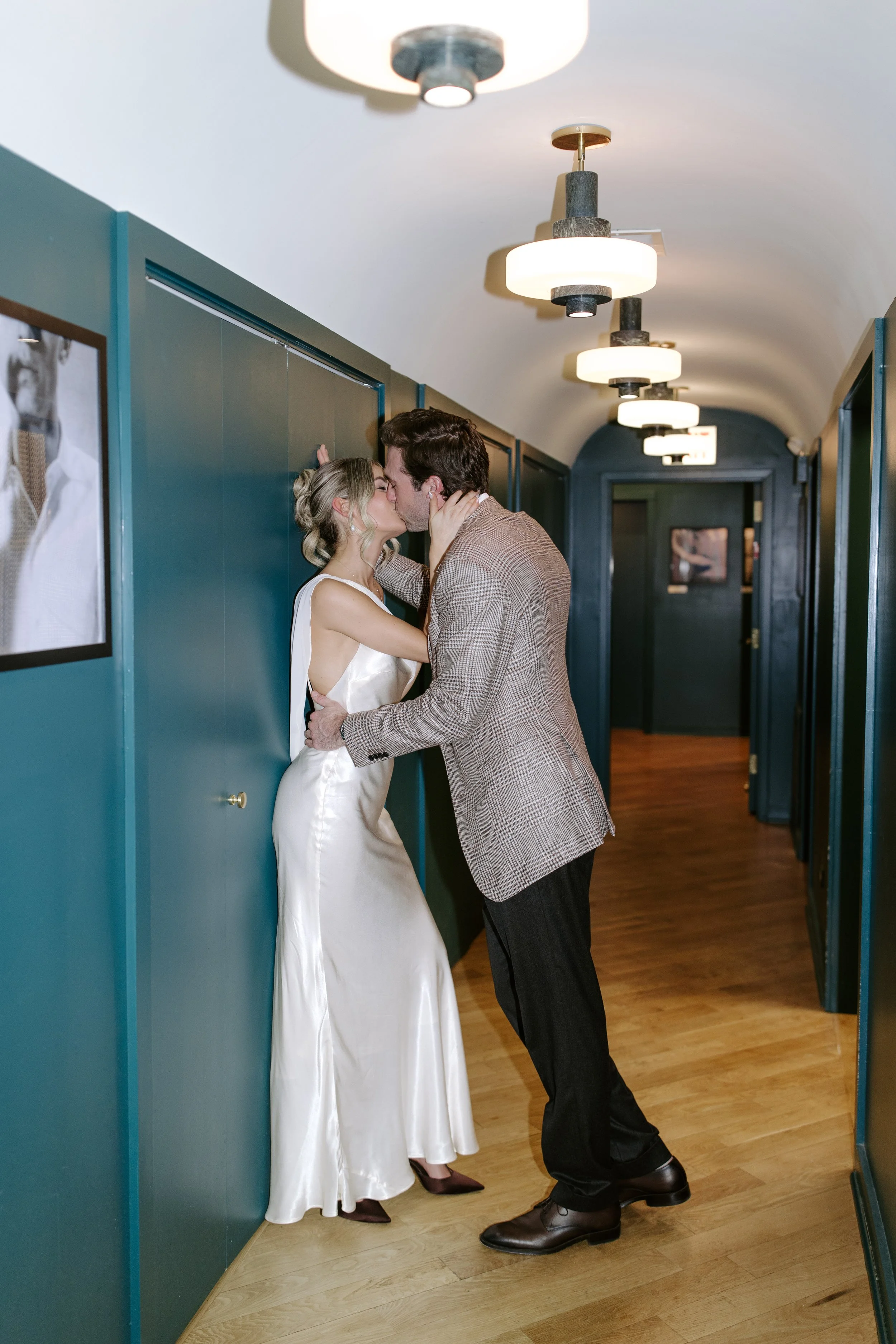 A man and woman share a kiss in a hallway with teal walls and modern lighting fixtures. The woman is wearing a white satin dress, and the man is dressed in a checkered blazer and dark pants.