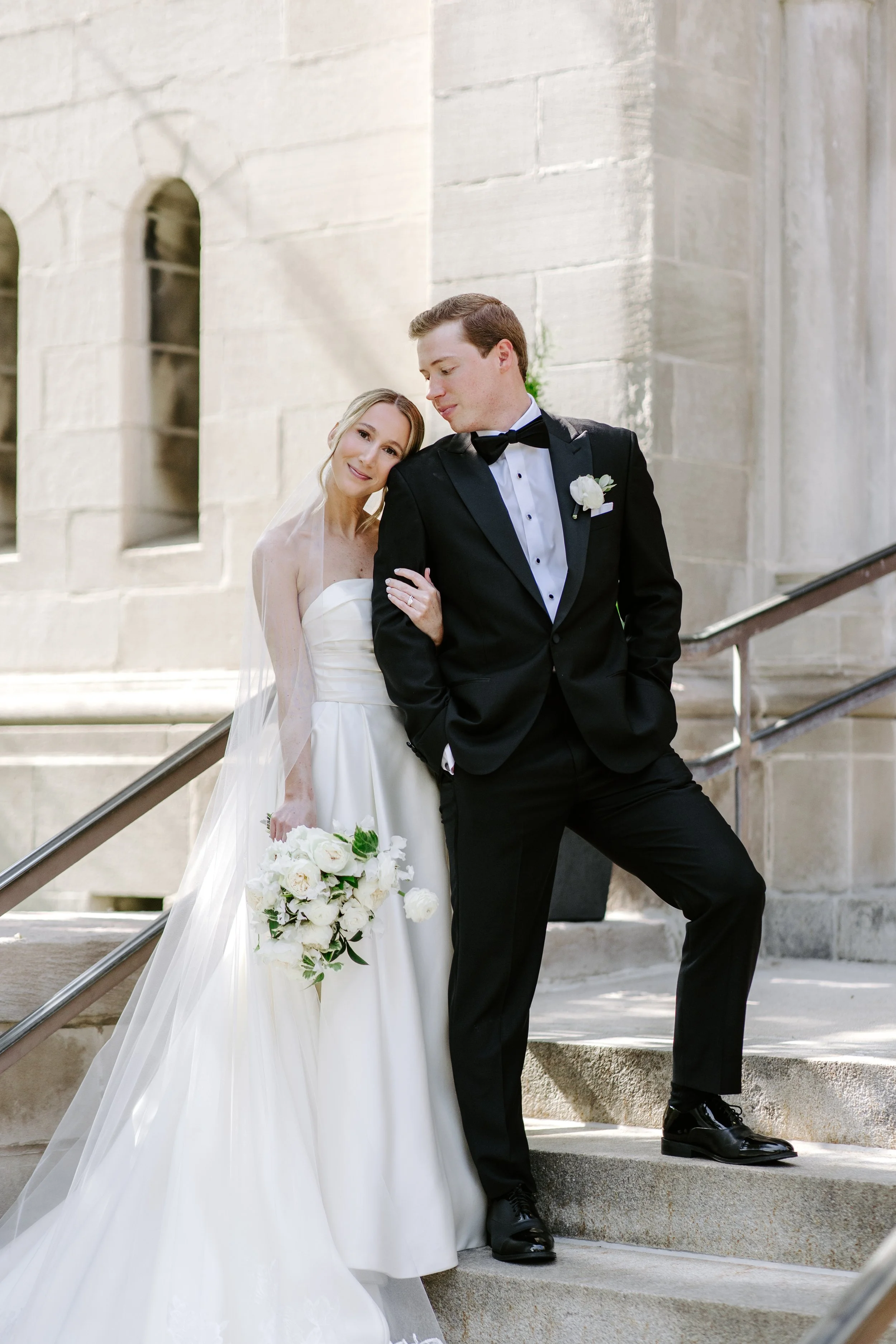 A bride and groom dressed in wedding attire standing on outdoor stone steps. The bride is holding a bouquet of white flowers and wearing a strapless white wedding gown with a veil, while the groom is wearing a black tuxedo with a bow tie and boutonni
