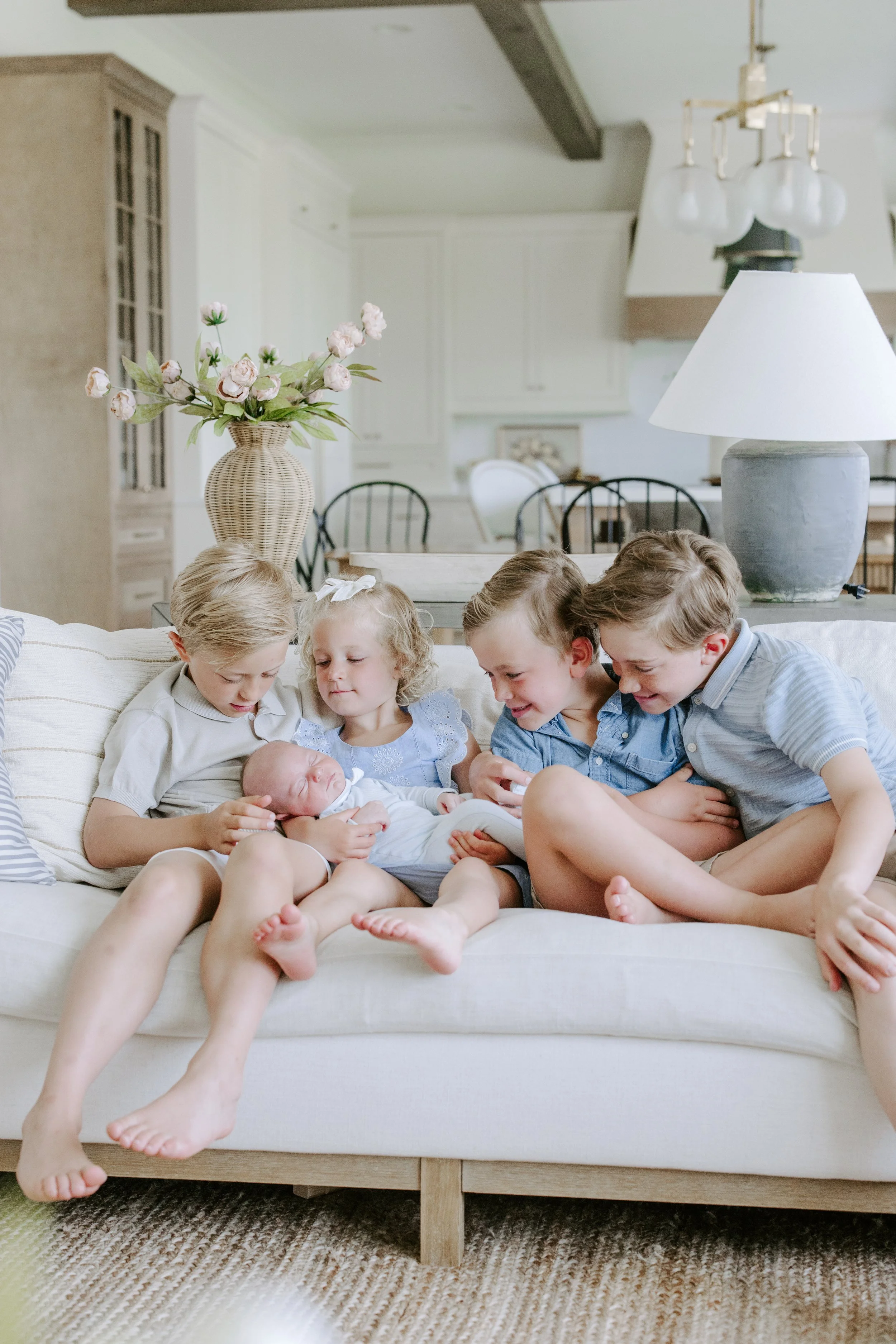 Five children sitting together on a white sofa in a living room, looking at a baby.
