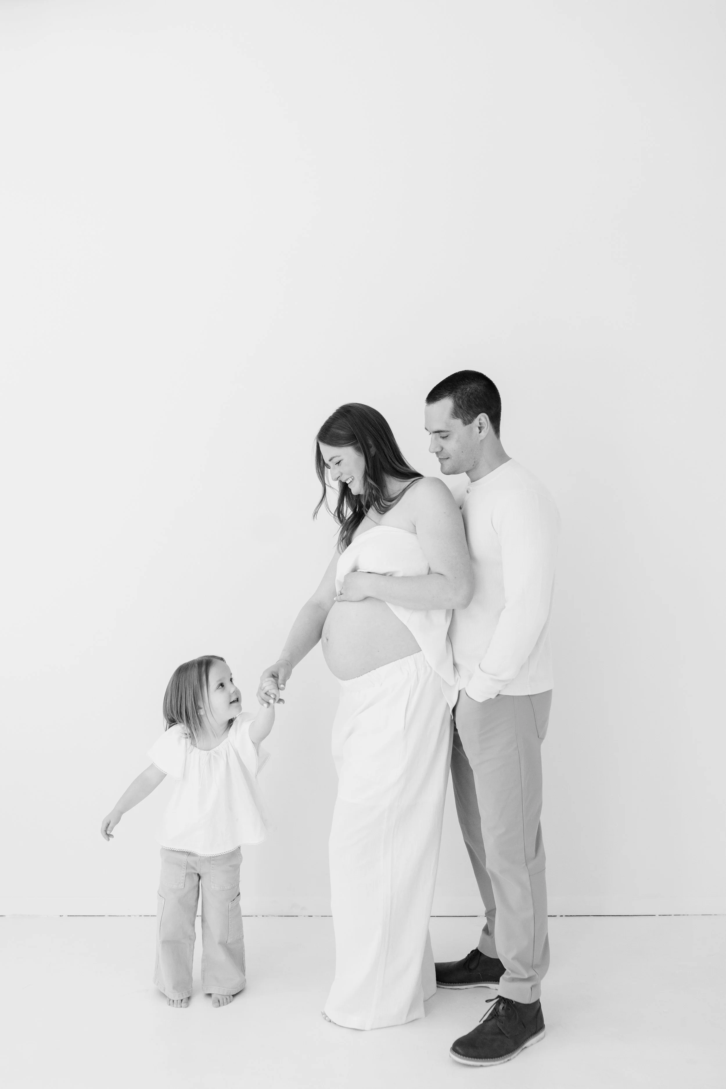 A black and white photograph of a happy family of four with a pregnant woman, her partner, and two young daughters, standing against a plain white background.