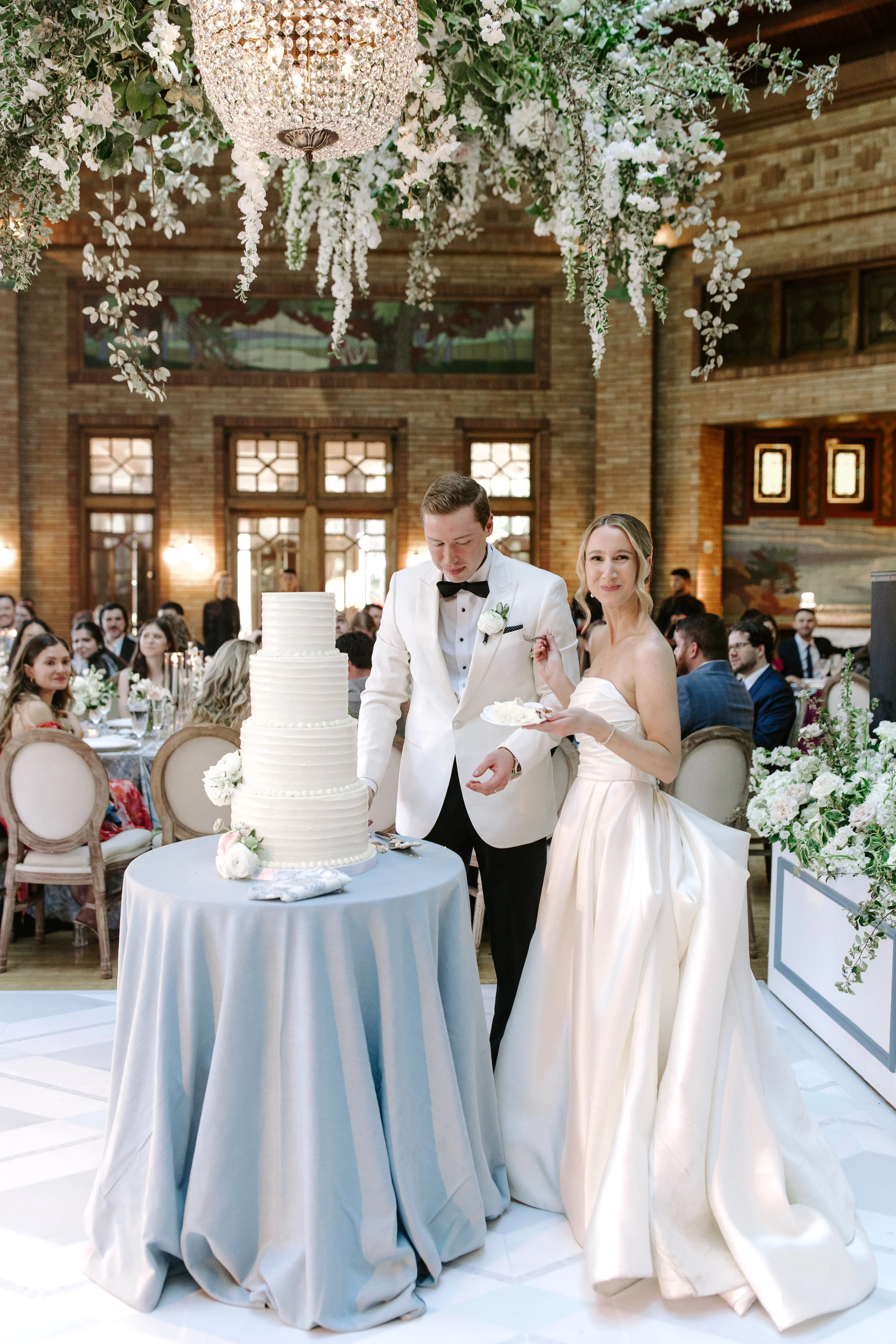A bride and groom cutting a white wedding cake decorated with flowers at their wedding reception in a decorated venue with floral arrangements and guests seated at tables.