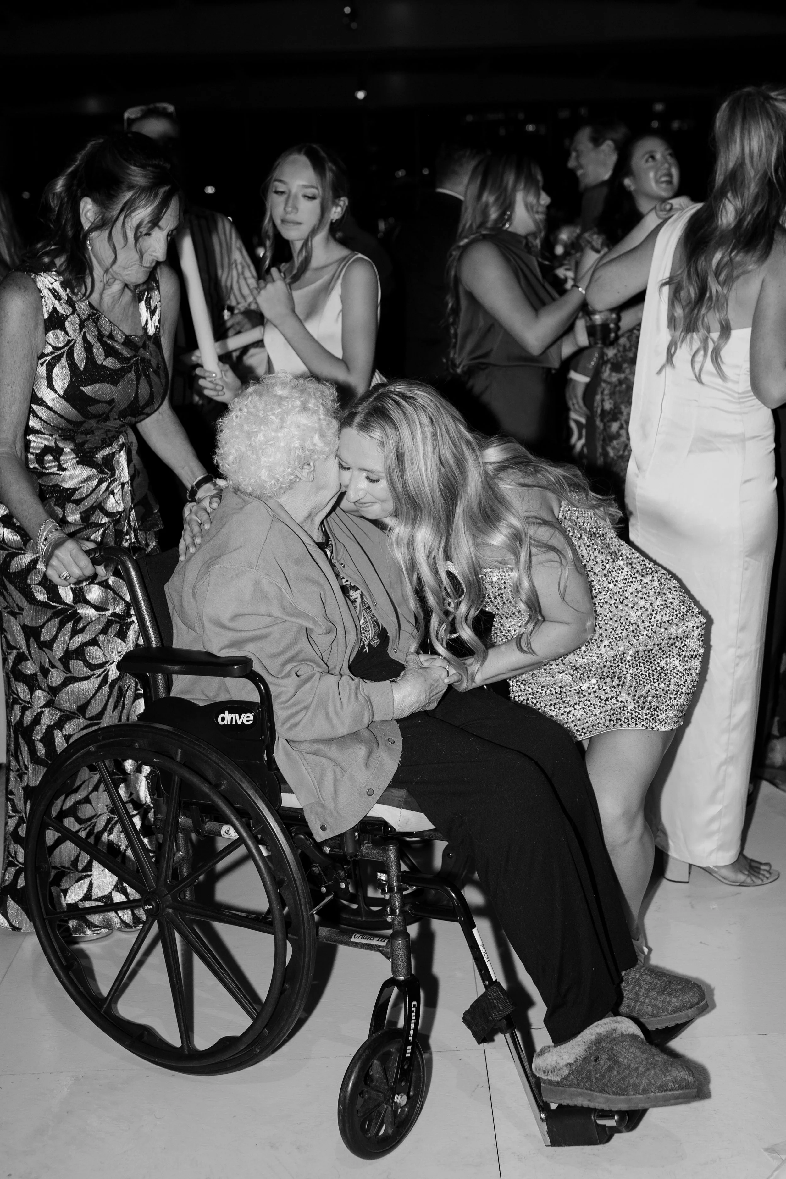 A woman in a wheelchair bends forward, holding hands with an elderly woman, in a social event with people dancing and talking in the background.