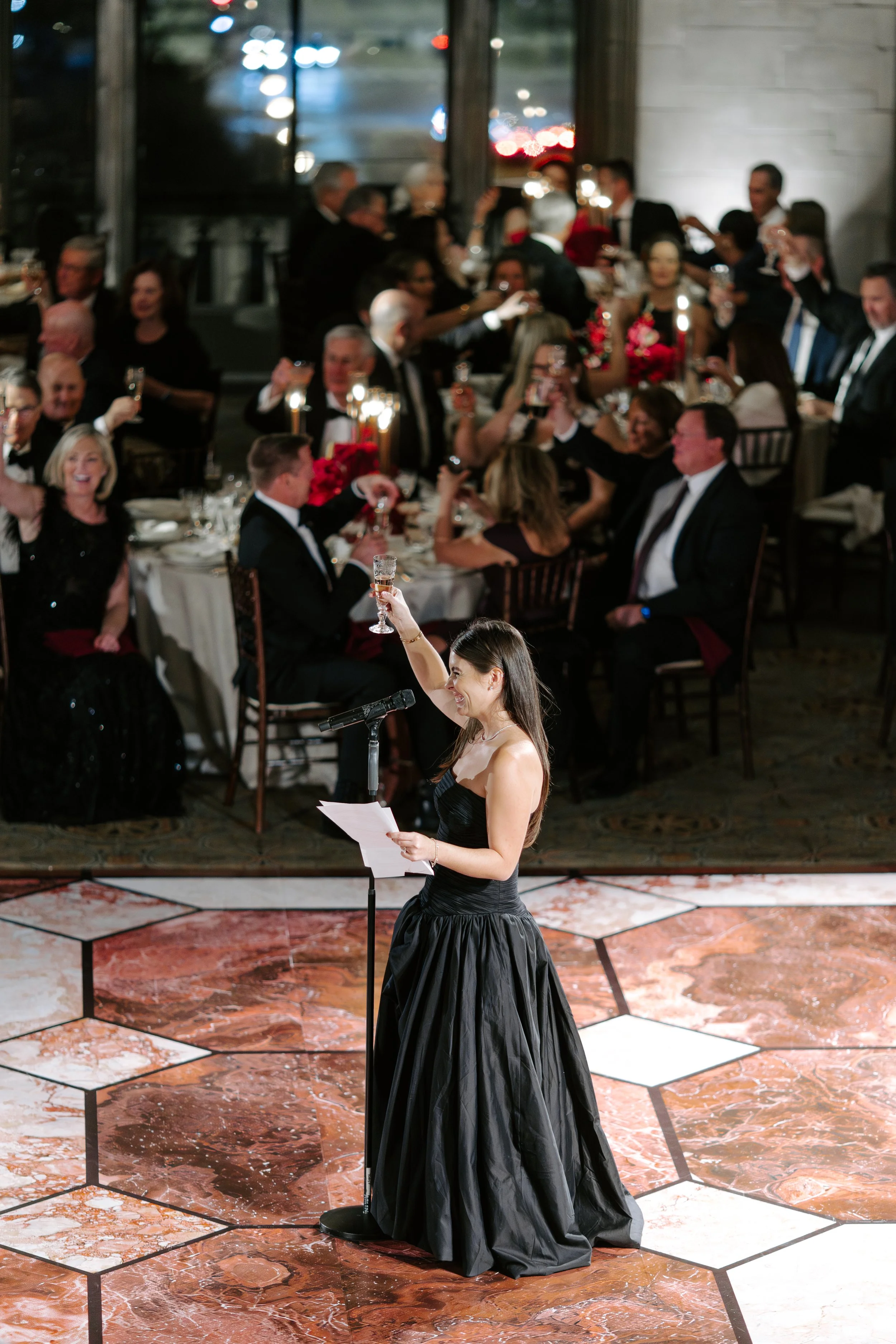 A woman in a black strapless gown giving a speech at a formal event or wedding reception, with a large group of people seated at decorated tables in the background, raising glasses for a toast.