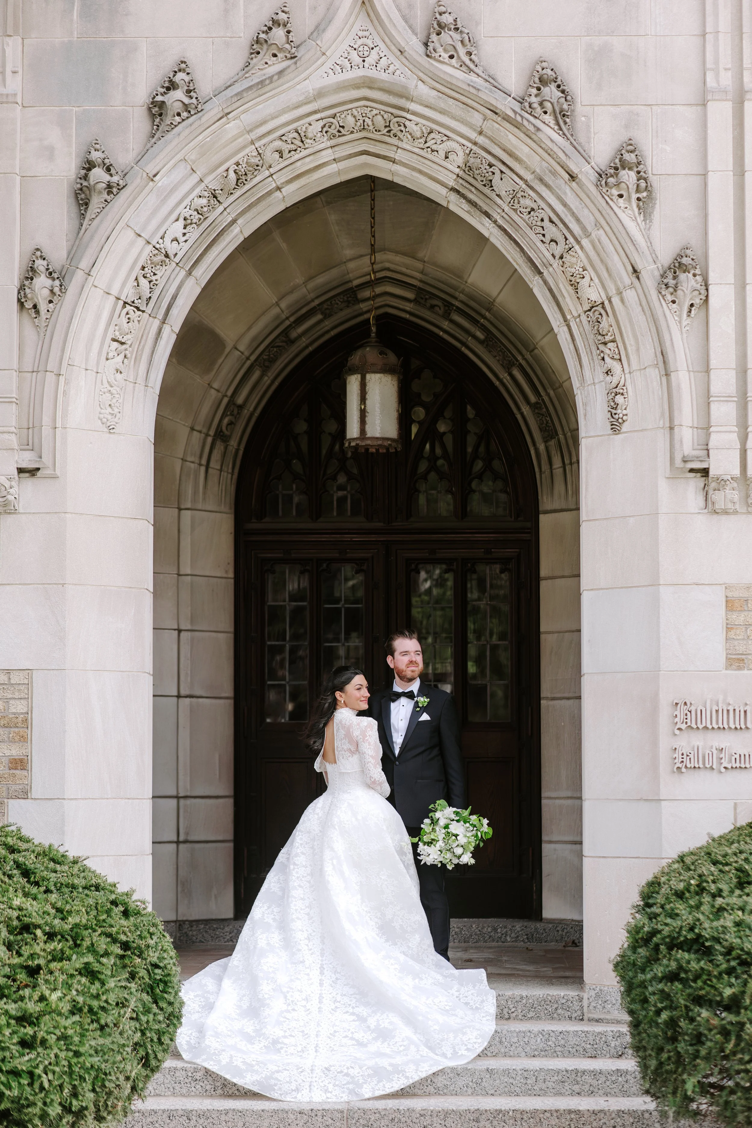 A bride and groom standing on the steps outside a building with arched doorway, intricate stone carvings, and a hanging lantern. The bride is in a white gown with lace long sleeves and a long train holding a bouquet of white flowers with greenery, an