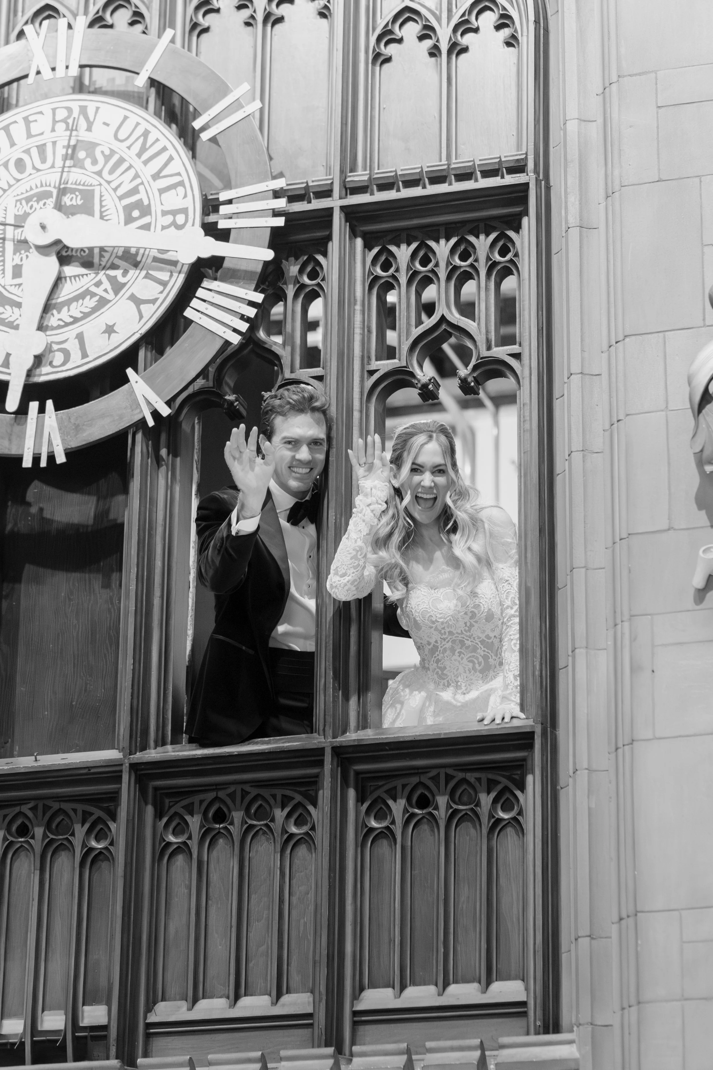 Two people, a man in a tuxedo and a woman in a wedding dress, are standing in a decorative wooden booth in front of a large clock face and Gothic architectural elements.