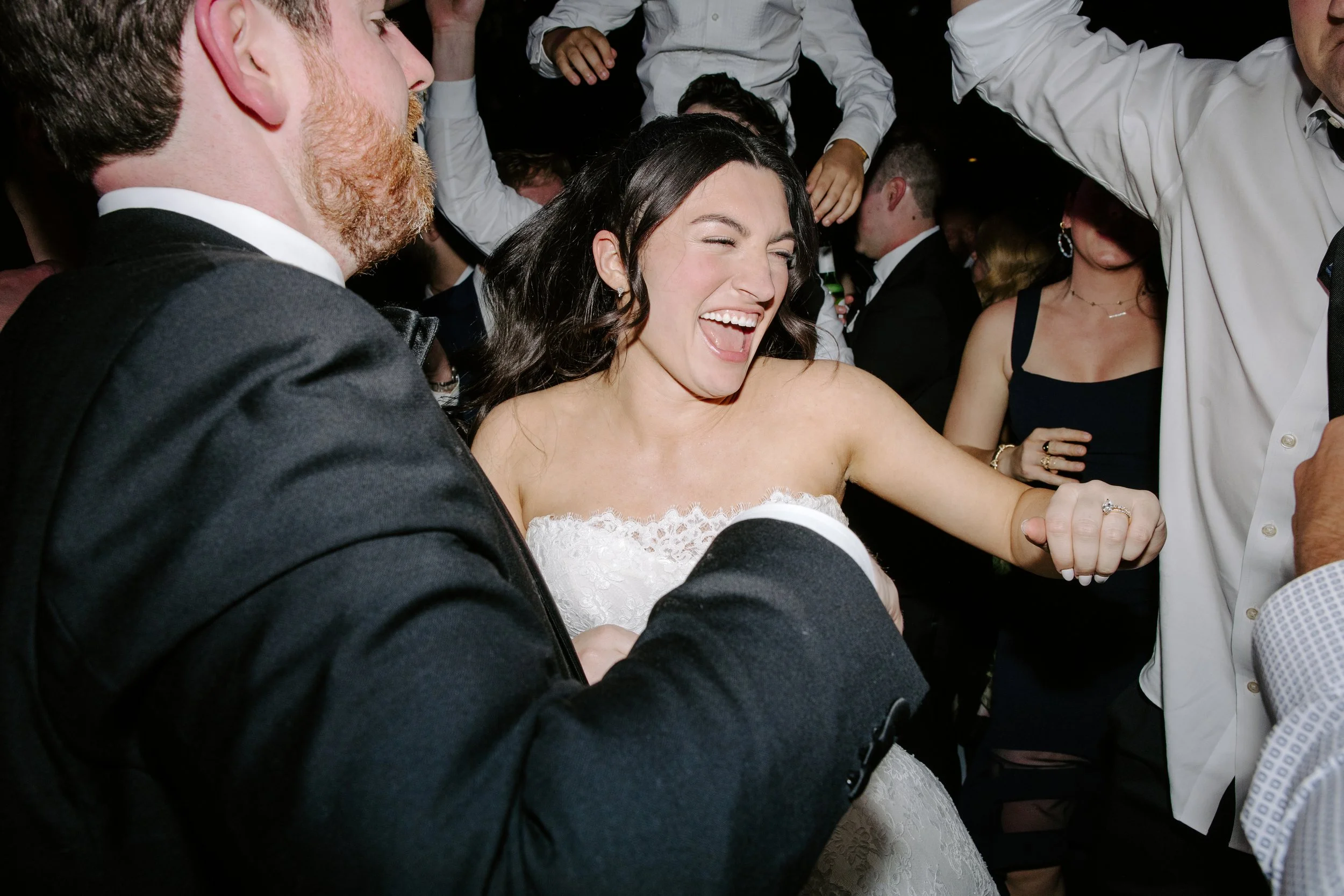 A bride in a white wedding dress laughing and dancing at her wedding reception, surrounded by wedding guests.